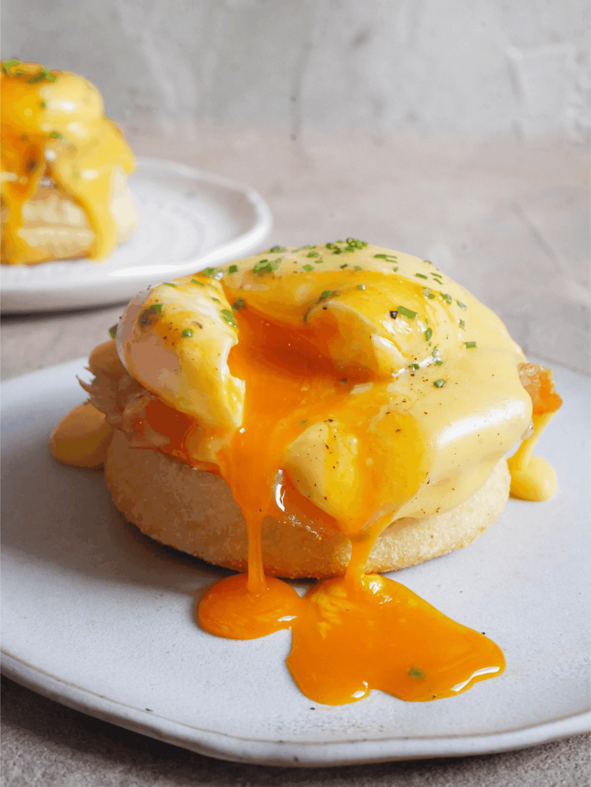 Eggs Benedict sits on white plate and poached egg has been cracked. Yolk ozzes out onto plate. Freshly cut chives and black pepper is sprinkled on top of eggs. 