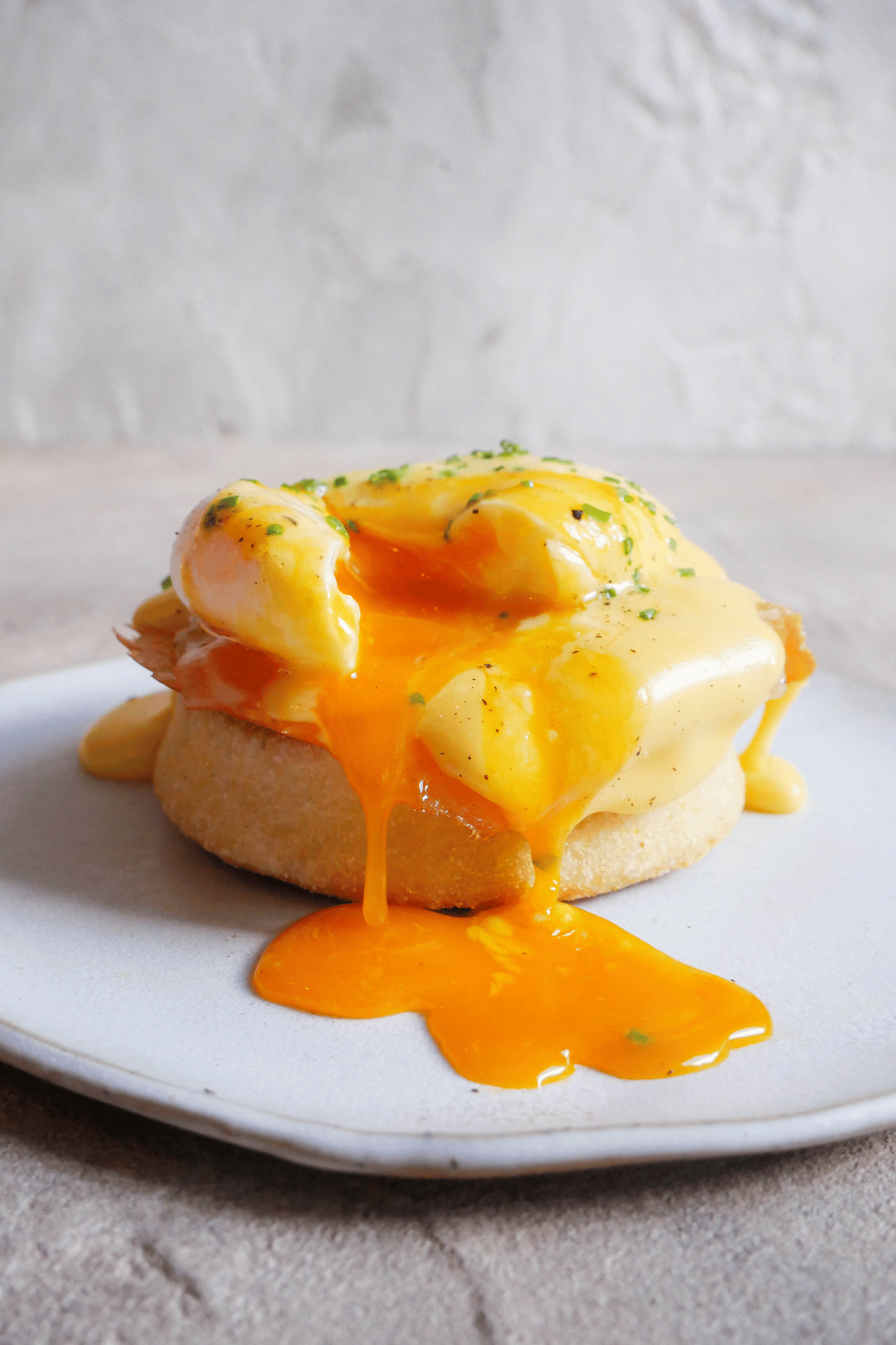 Eggs Benedict sits on white plate and poached egg has been cracked. Yolk ozzes out onto plate. Freshly cut chives and black pepper is sprinkled on top of eggs.