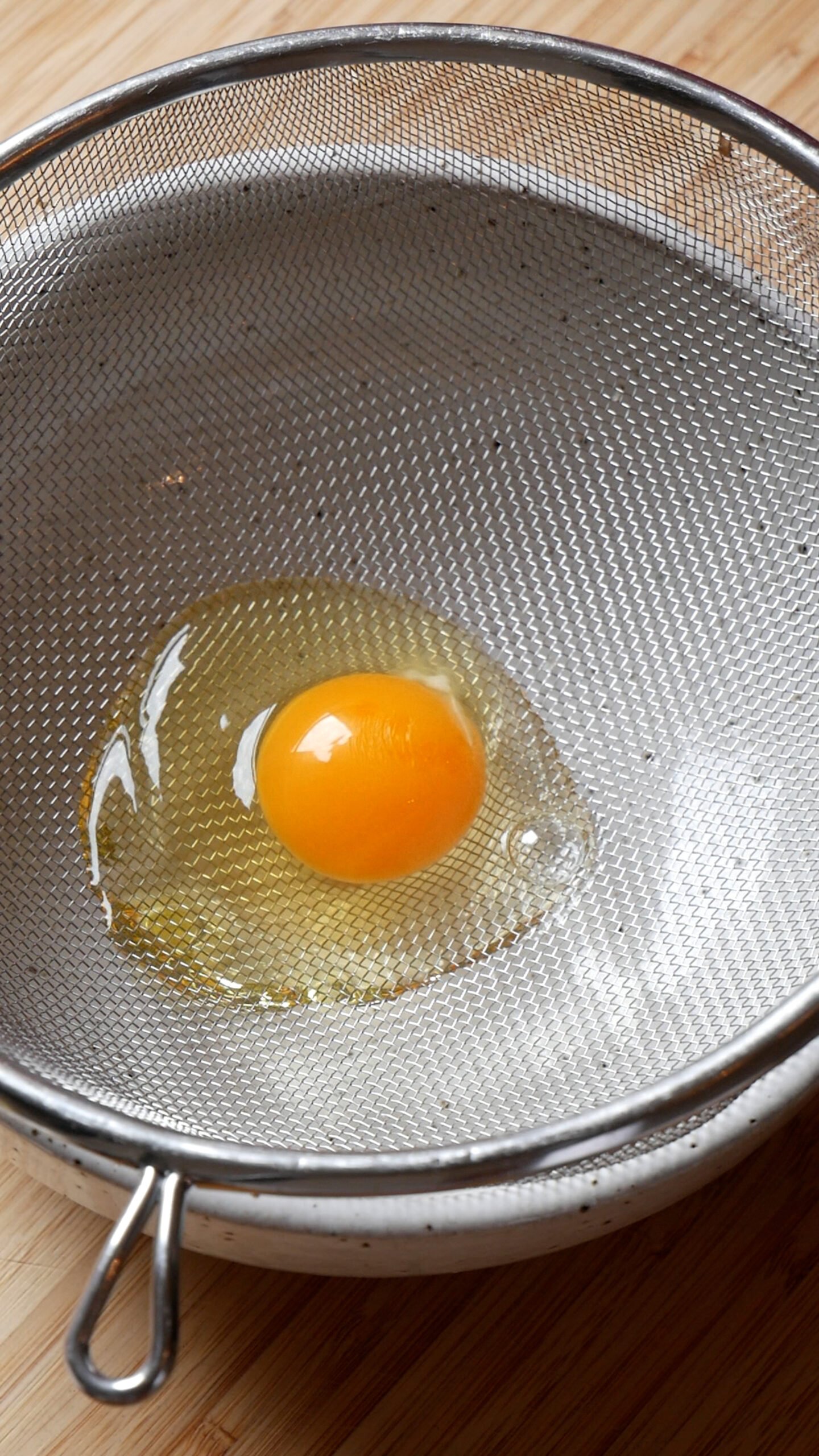 Cracked egg sits in fine mesh strainer sitting over white bowl. 