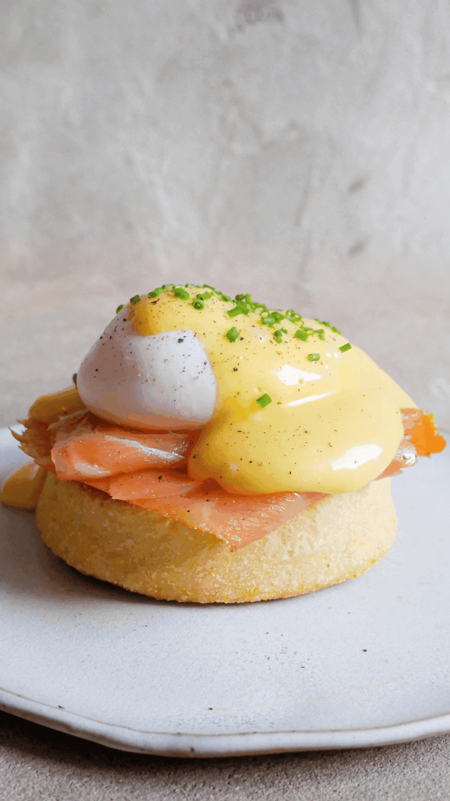 Eggs benedict sit on white plate. You can see english muffin, smoked salmon, egg on top with hollandaise and chives and pepper. 