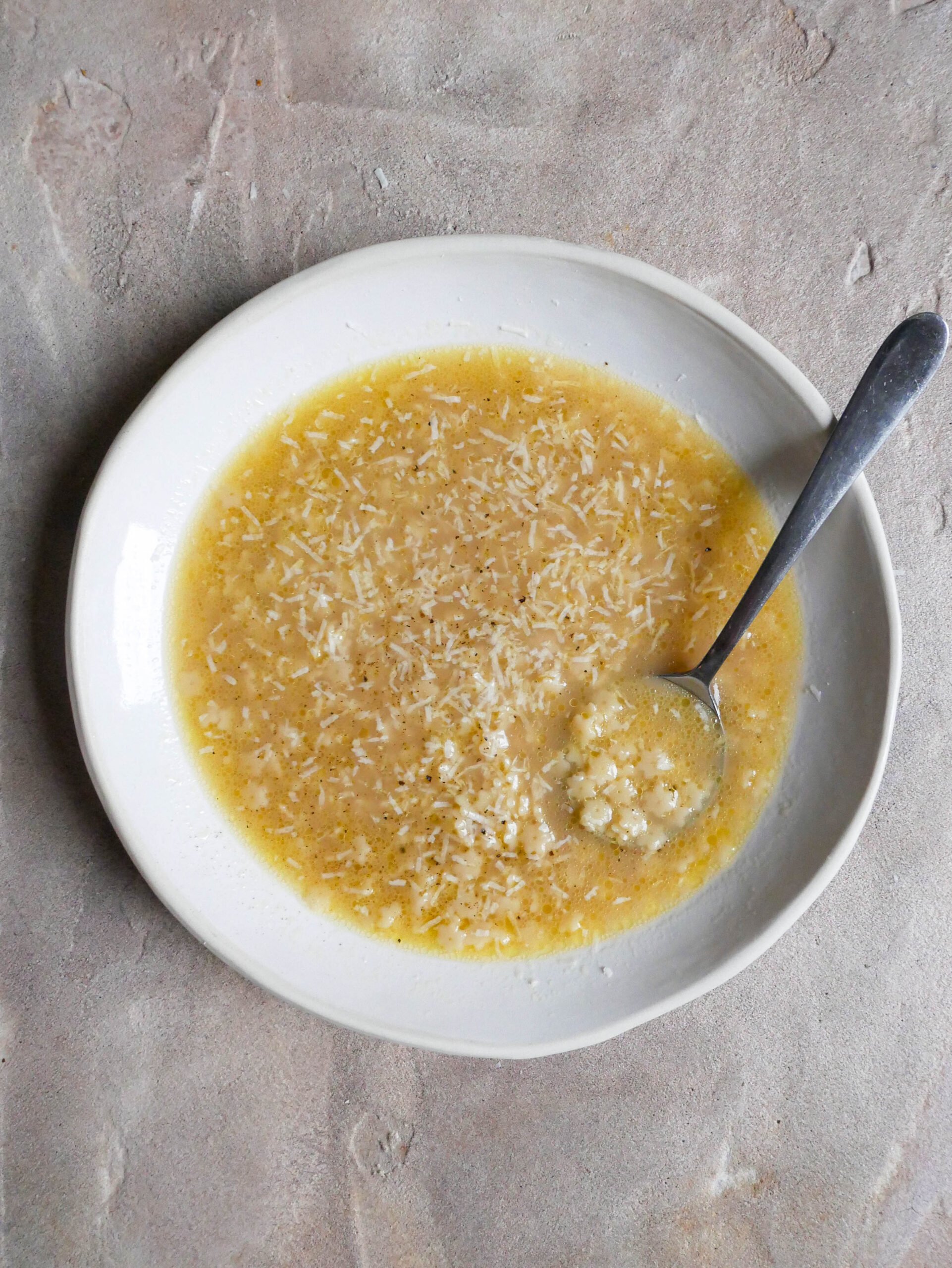 Pastina in chicken broth in a bowl. There is a bowl sticking out the side of the bowl and grated cheese is on top.