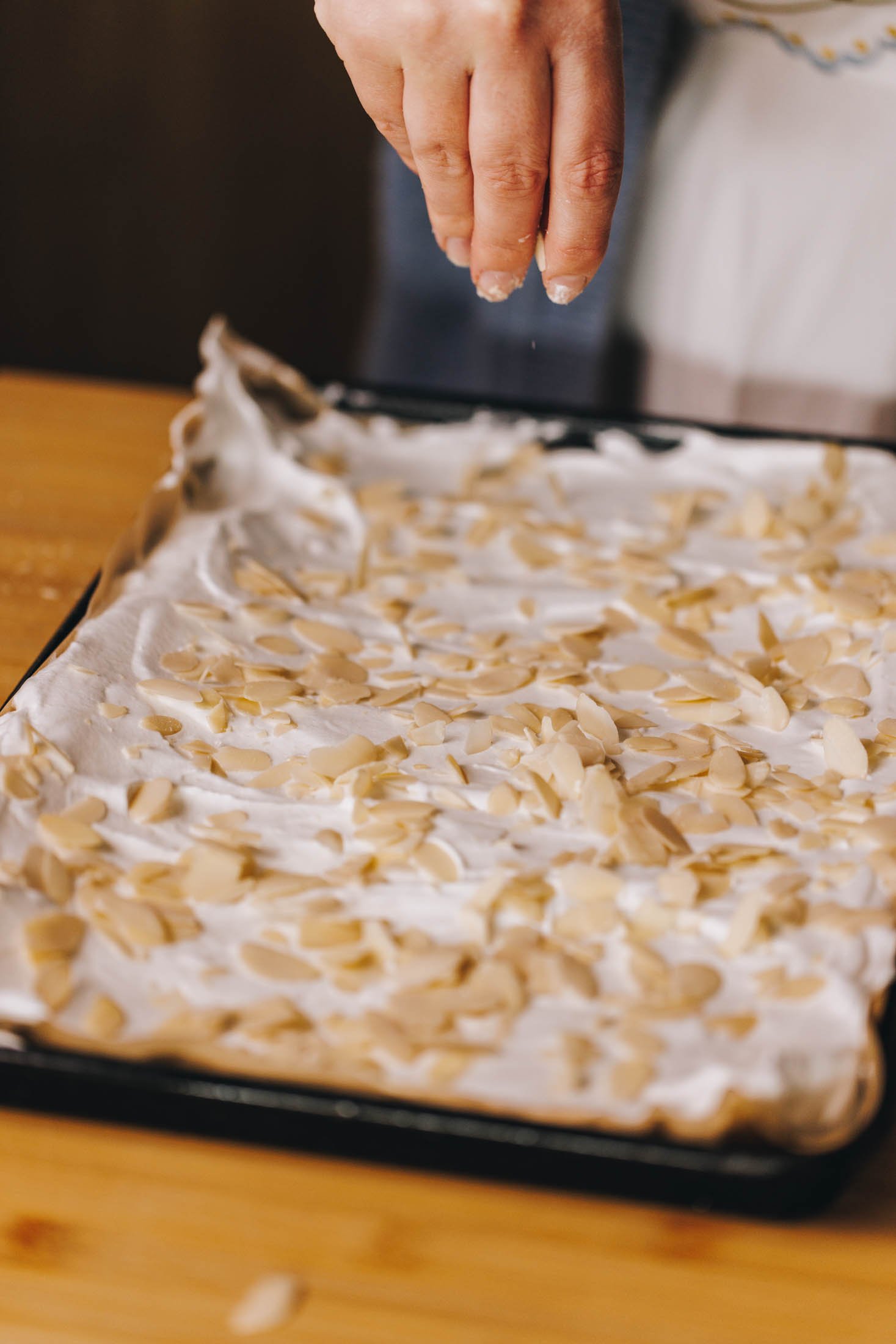 Slithered almonds are being sprinkled onto meringue on baking tray.