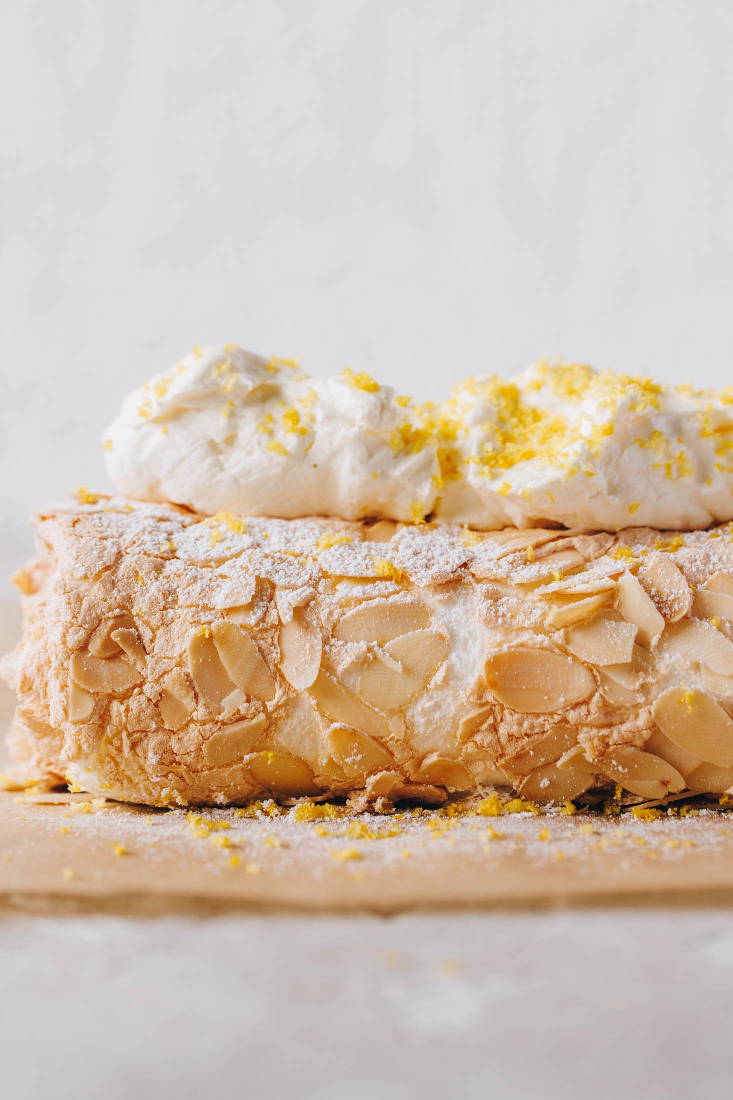 Lemon curd meringue roulade sits on baking paper and has whipped cream and lemon zest on top.
