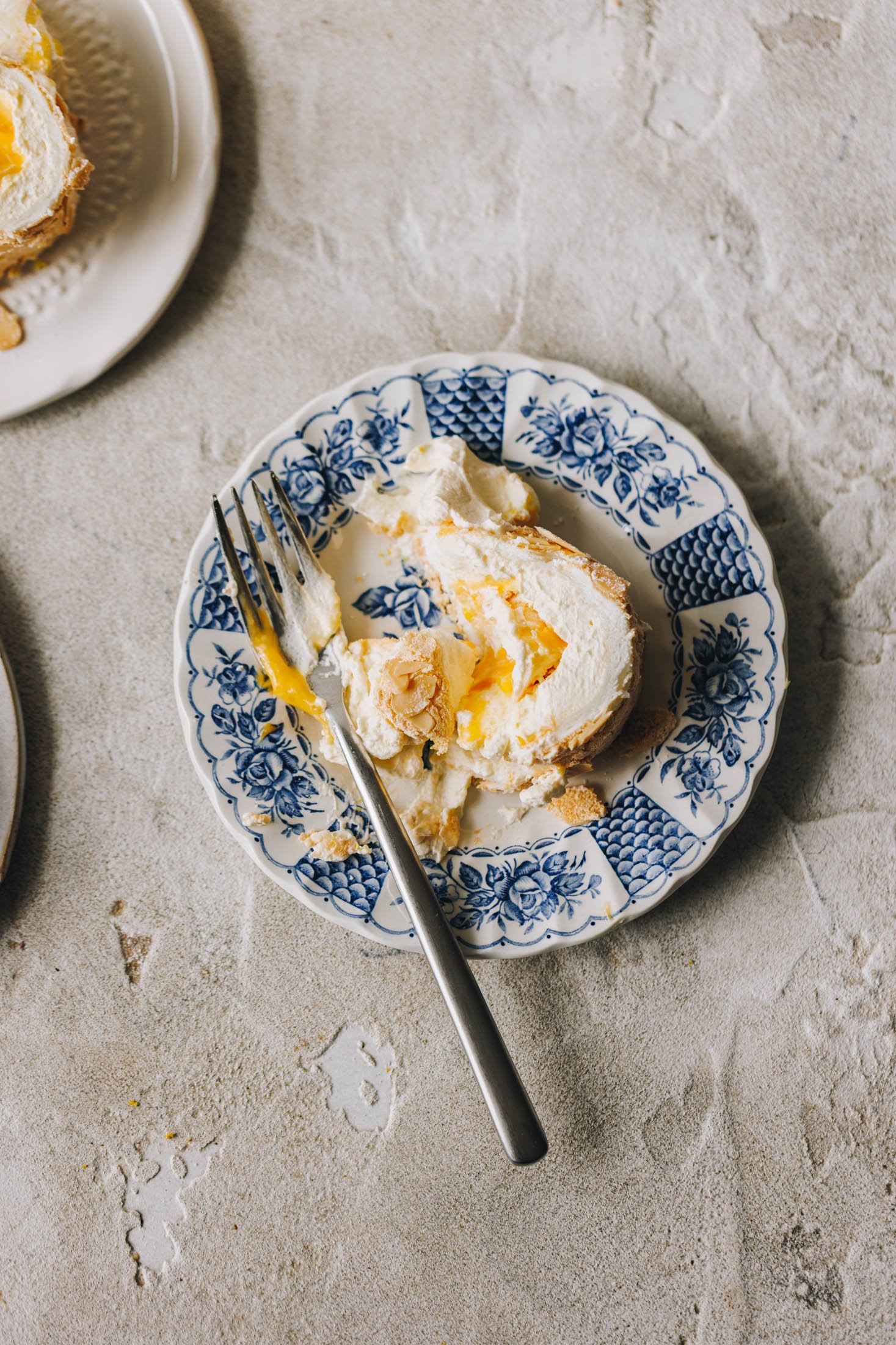 A slice of lemon curd meringue roulade sits on a blue and white plate with a floral pattern. A slice has been taken out of it with a fork.