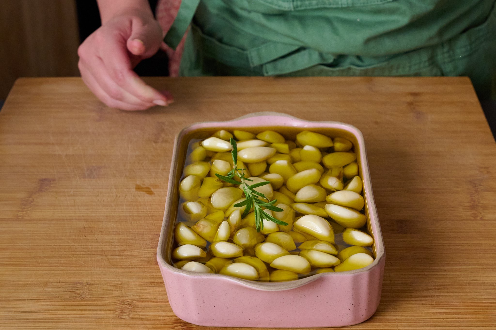 Garlic are in a pink baking dish and filled with olive oil. There is a sprig of rosemary.