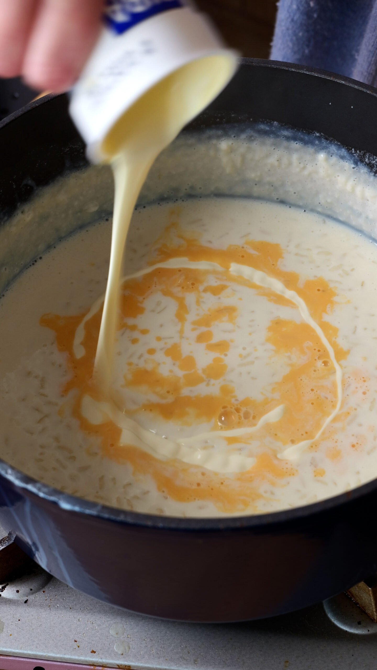 Egg yolks and cream are being poured into the cooked rice and milk in a large blue pot.