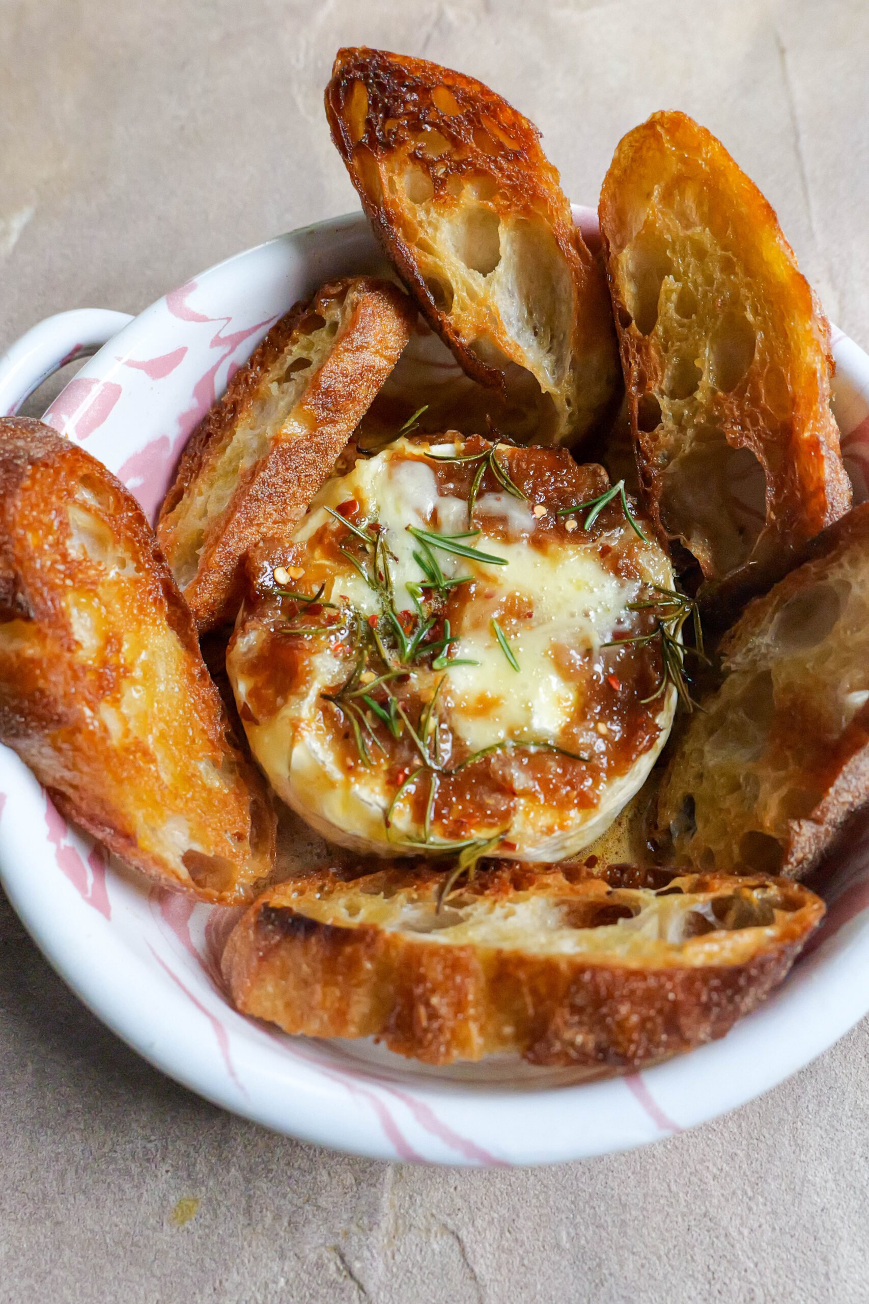 Baked brie with roasted garlic butter sits in pink and white tray with olive oil fried slices of baguette