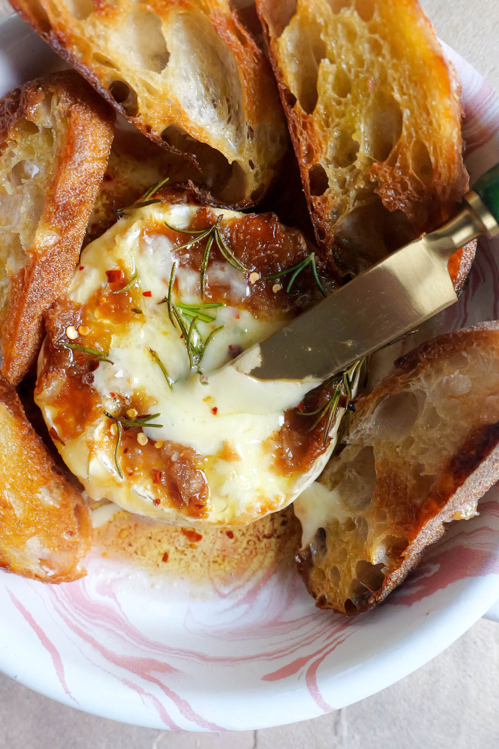 Baked brie with roasted garlic butter sits in pink and white tray with olive oil fried slices of baguette. There is also a knife sticking out of the brie. 