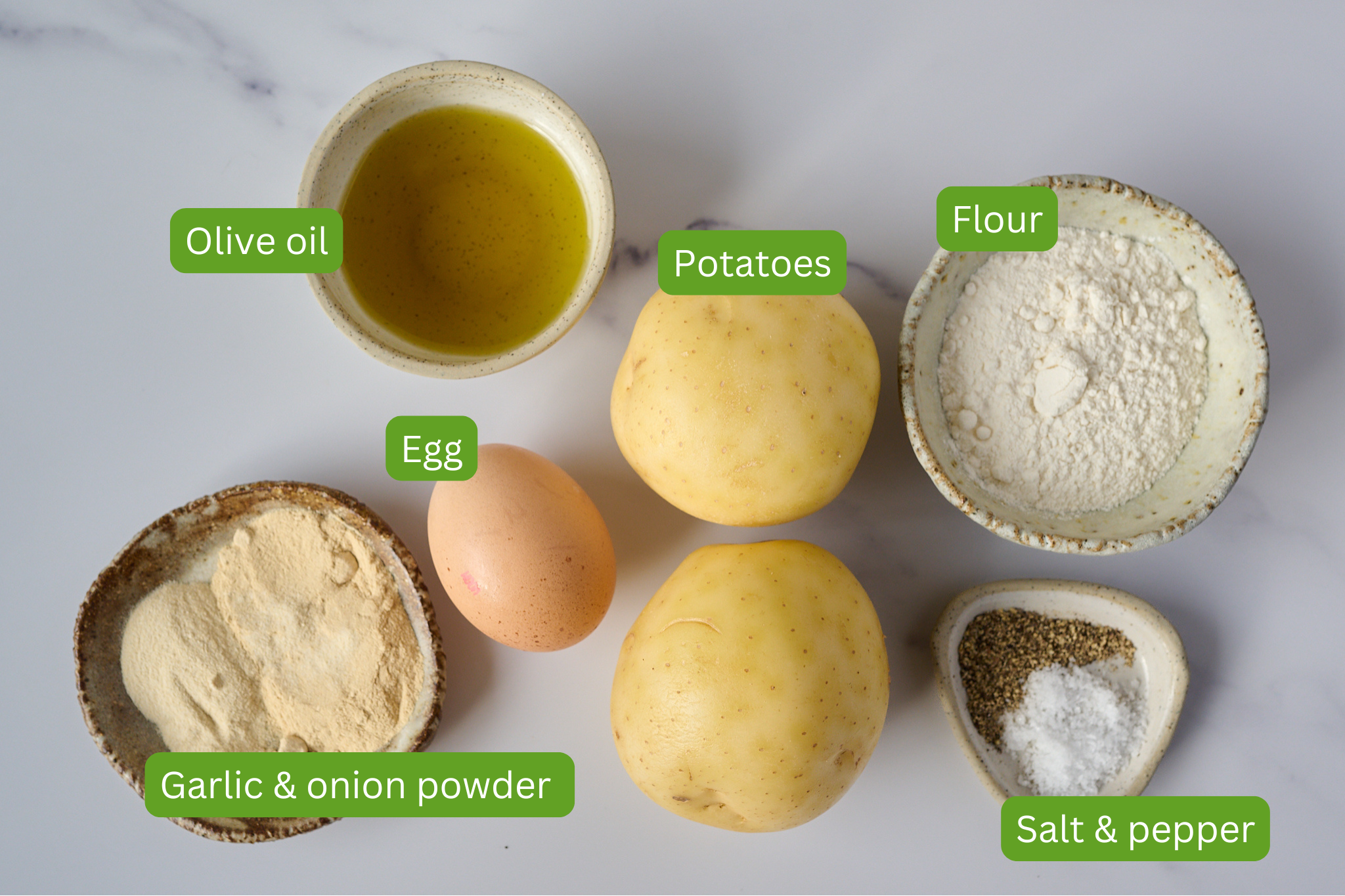 List of ingredients are laid out for potato rosti on a white marble backdrop. Ingredients include potato, flour, garlic and onion powder, salt and pepper, egg and olive oil. 
