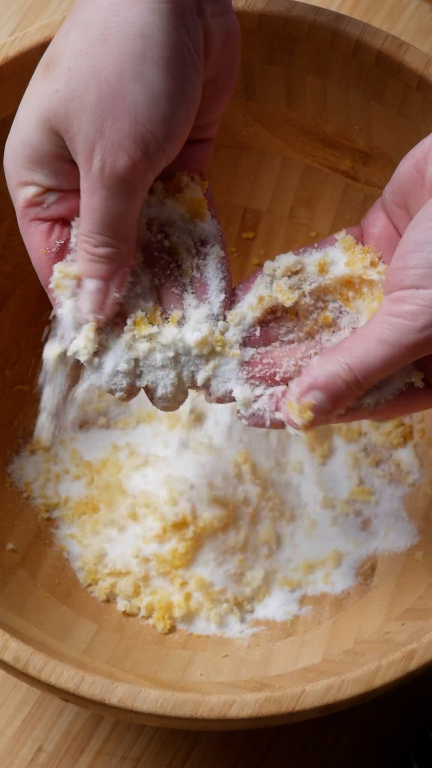 Hands are rubbing lemon zest and sugar together over a large wooden bowl.