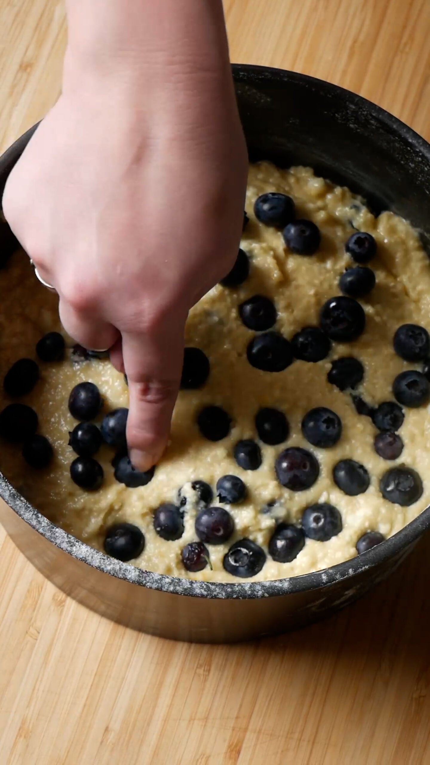 Cake batter is in black cake tin and finger is poking bluberries into batter.