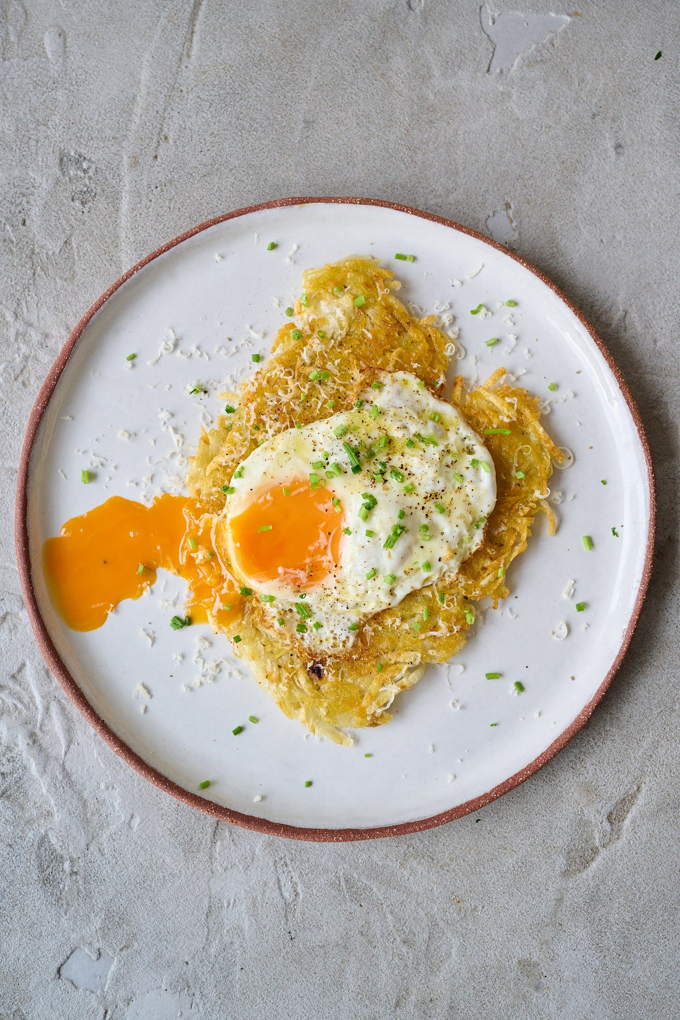Potato rosti sits on a white plate with a brown rim. It has a fried egg that has been broken and the yolk oozing out sitting on top. You can see finely cut chives and parmesan sprinkled on top.