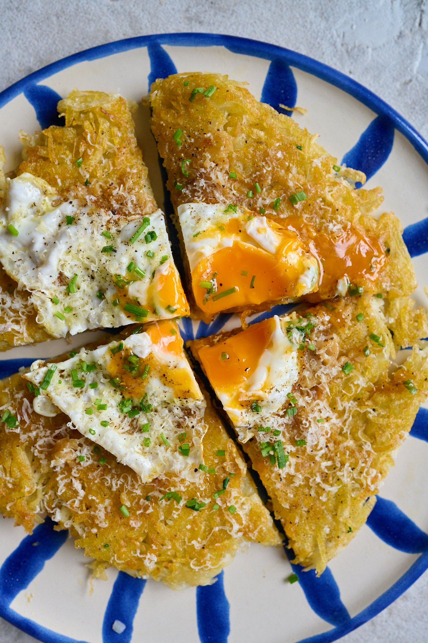 potato rosti is cut into fours and has a fried egg that has been broken sitting on it. The rosti sits on a white plate with blue lined. And the rosti has chives and cheese sprinkled all over. 