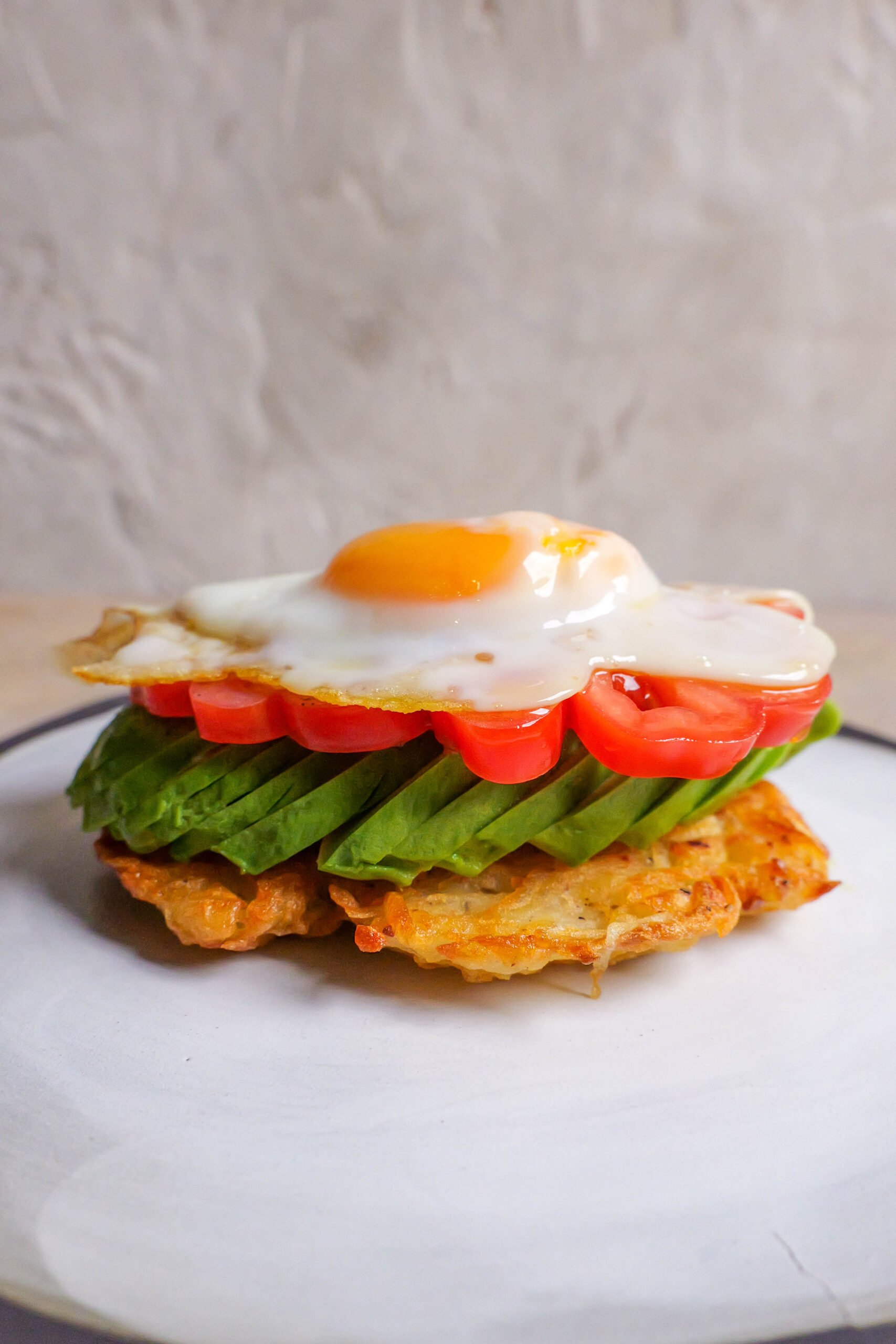 potato rosti sits on a white plate and has sliced avocado, tomato and fried egg sitting on top. 
