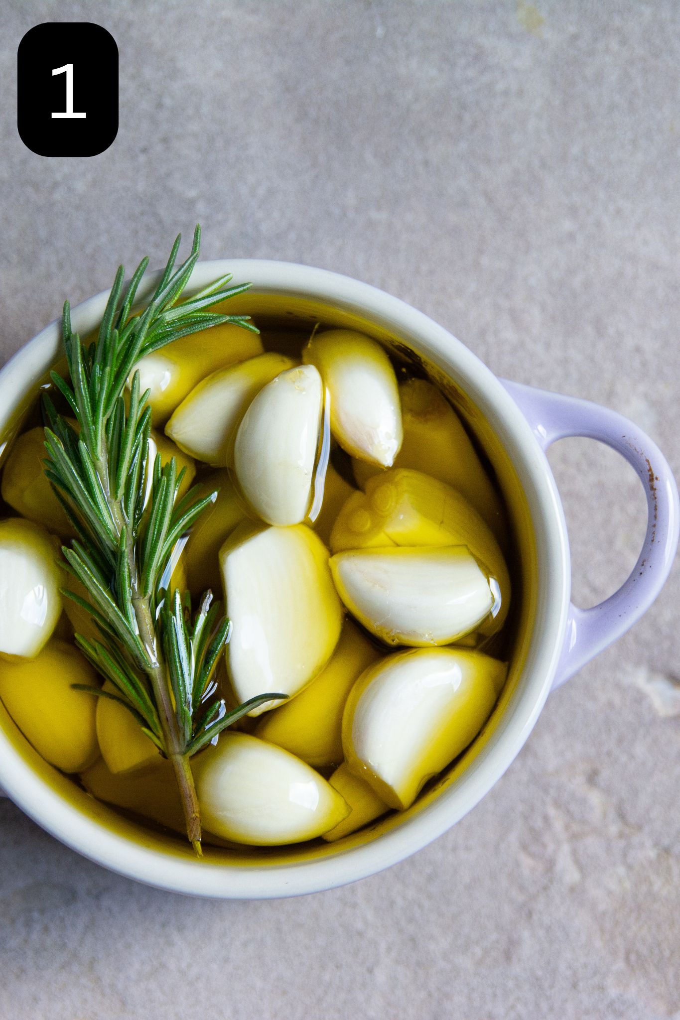 Garlic in smal purple bowl submerged in olive oil with rosemary sprig.