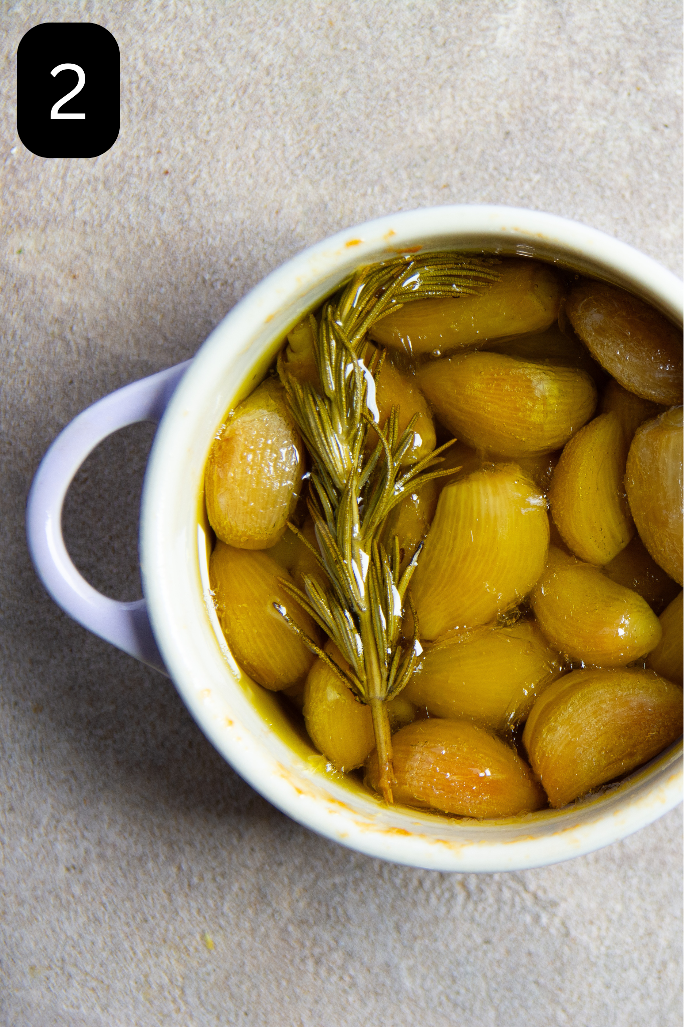 Cooked garlic in smal purple bowl submerged in olive oil with rosemary sprig.