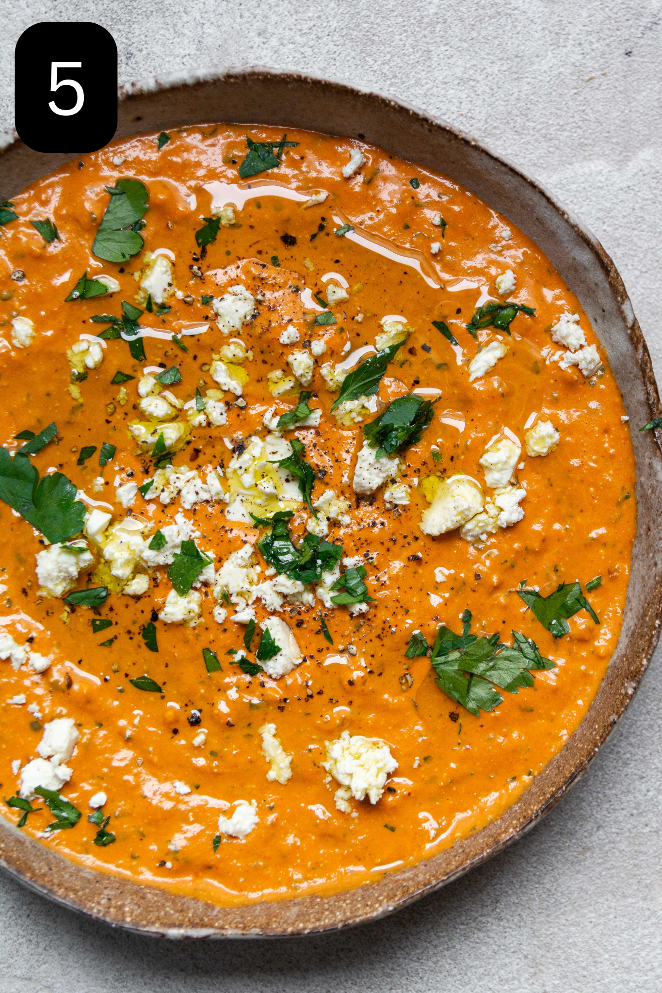 A large bowl filled with red pepper dip. Topped with feta, chopped parsley and a drizzle of olive oil