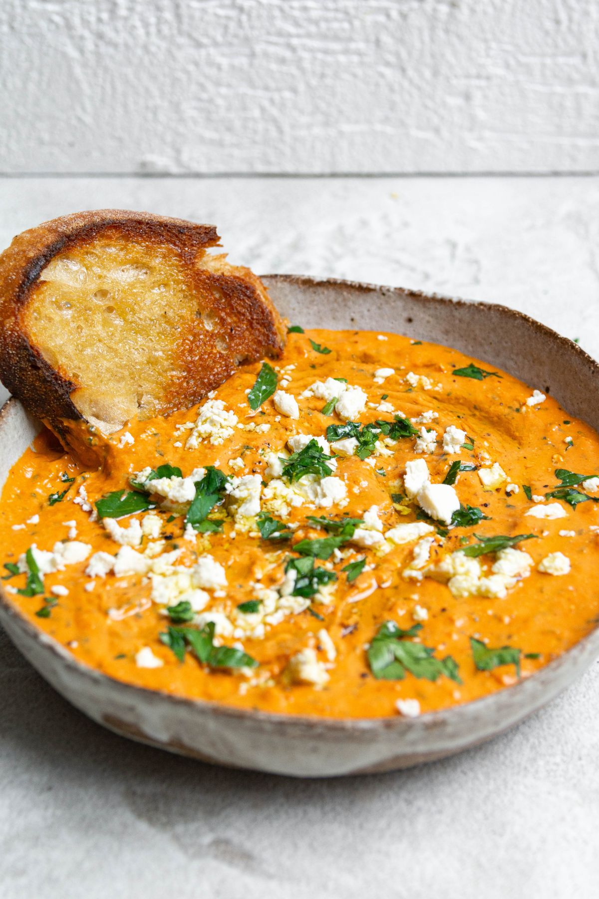 A large bowl filled with red pepper dip. Topped with feta, chopped parsley and a drizzle of olive oil. A piece of fried sourdough is poking out the side. 