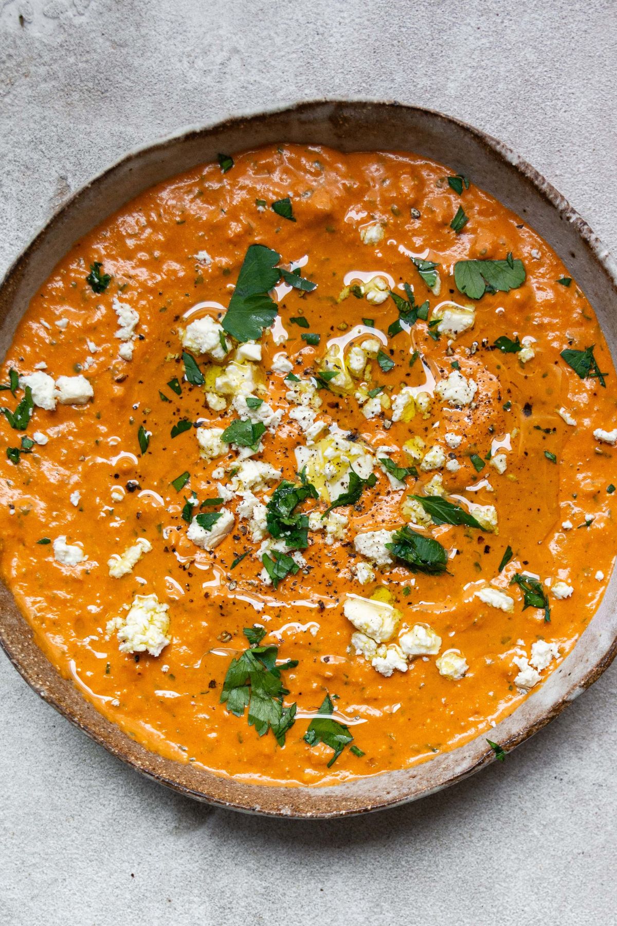 A large bowl filled with red pepper dip. Topped with feta, chopped parsley and a drizzle of olive oil