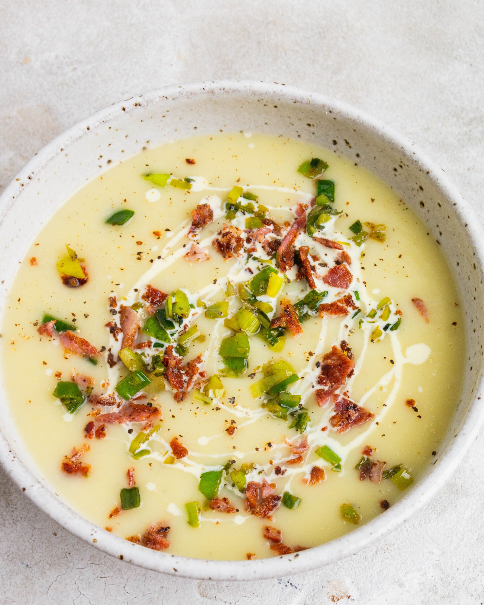 Potato and leek soup sits in large white bowl with leek tops and bacon garnished on top. There is also a drizzle of cream.