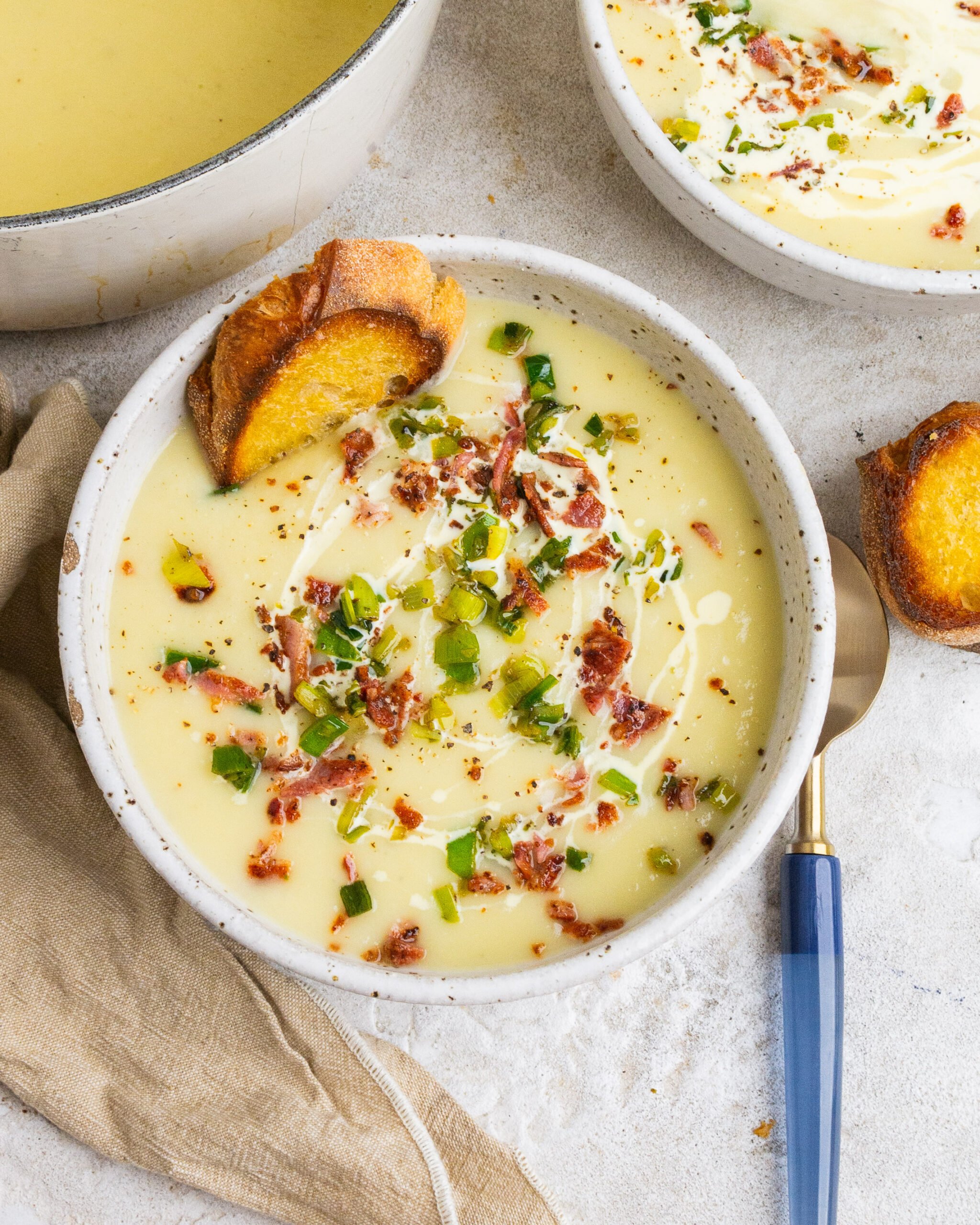 Potato and leek soup sits in large white bowl with leek tops and bacon garnished on top. There is also a drizzle of cream. Blue spoon is on right side of bowl. 