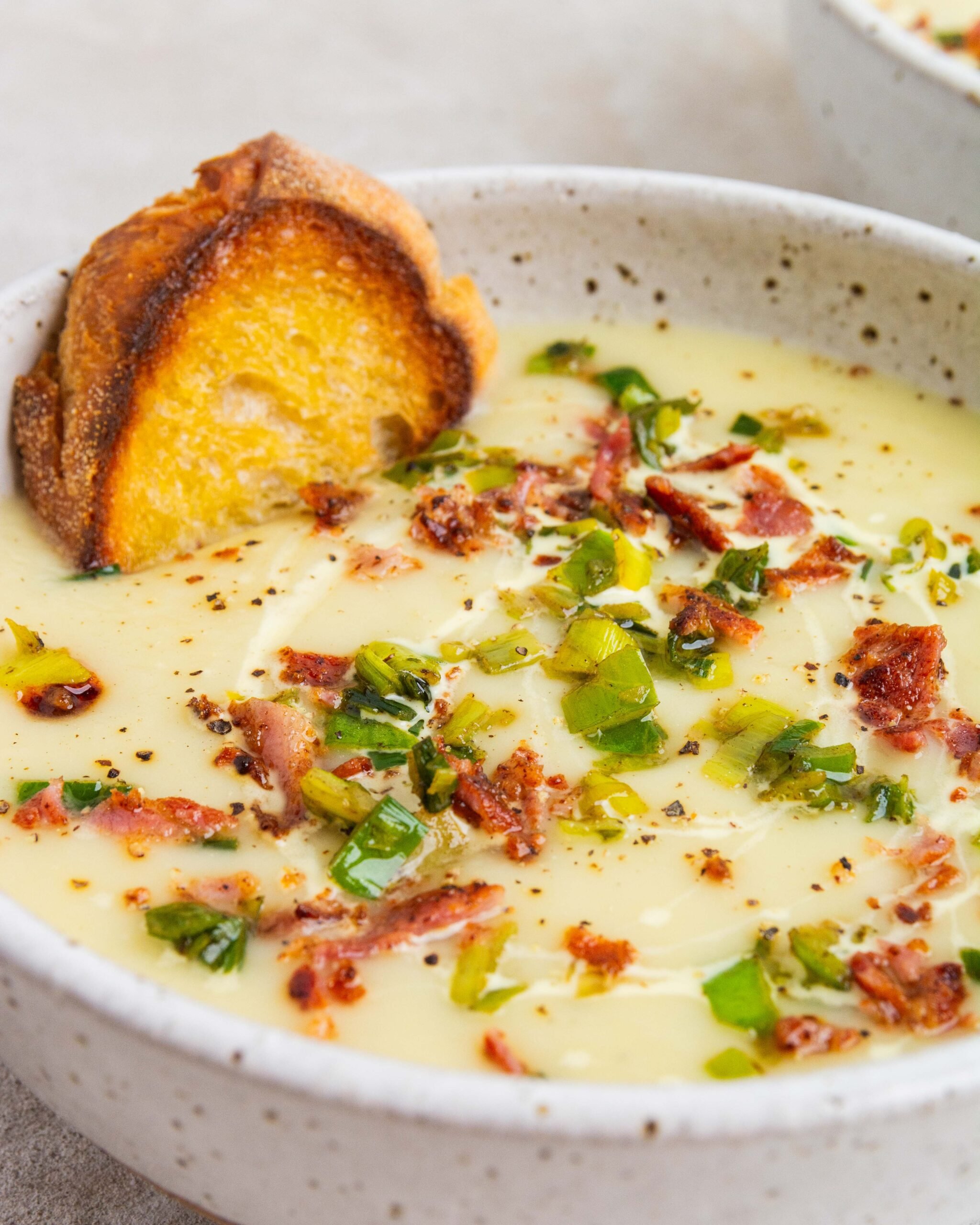 Potato and leek soup sits in large white bowl with leek tops and bacon garnished on top. There is also a drizzle of cream. Blue spoon is on right side of bowl.  Crispy bread is also sitting in bowl. 