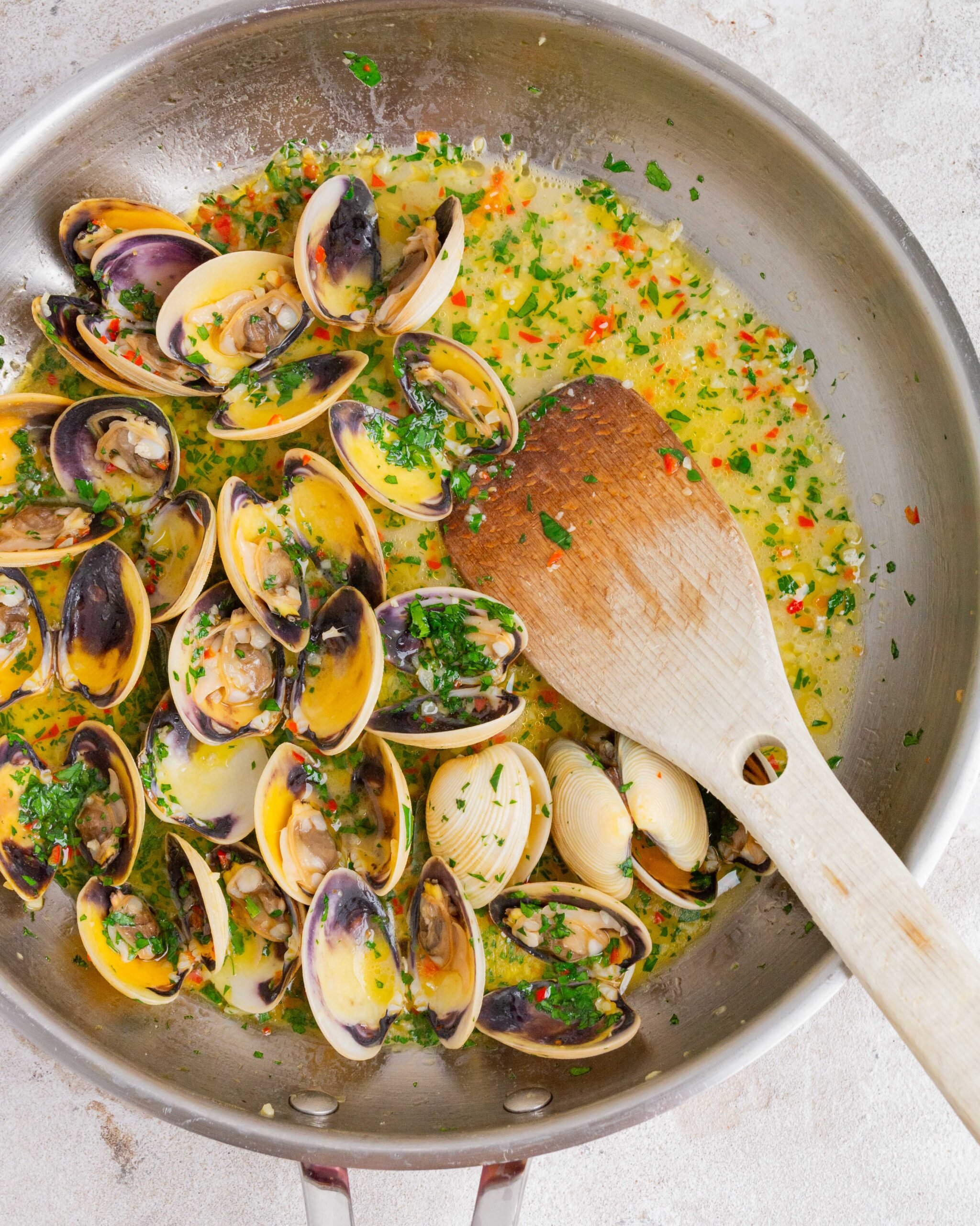 Garlic butter pipis / clams sit in large stainless steel pan with wooden spoon sticking out the right side. They have parsley sprinkled over them and you can see lots of minced garlic and sliced chilli. 