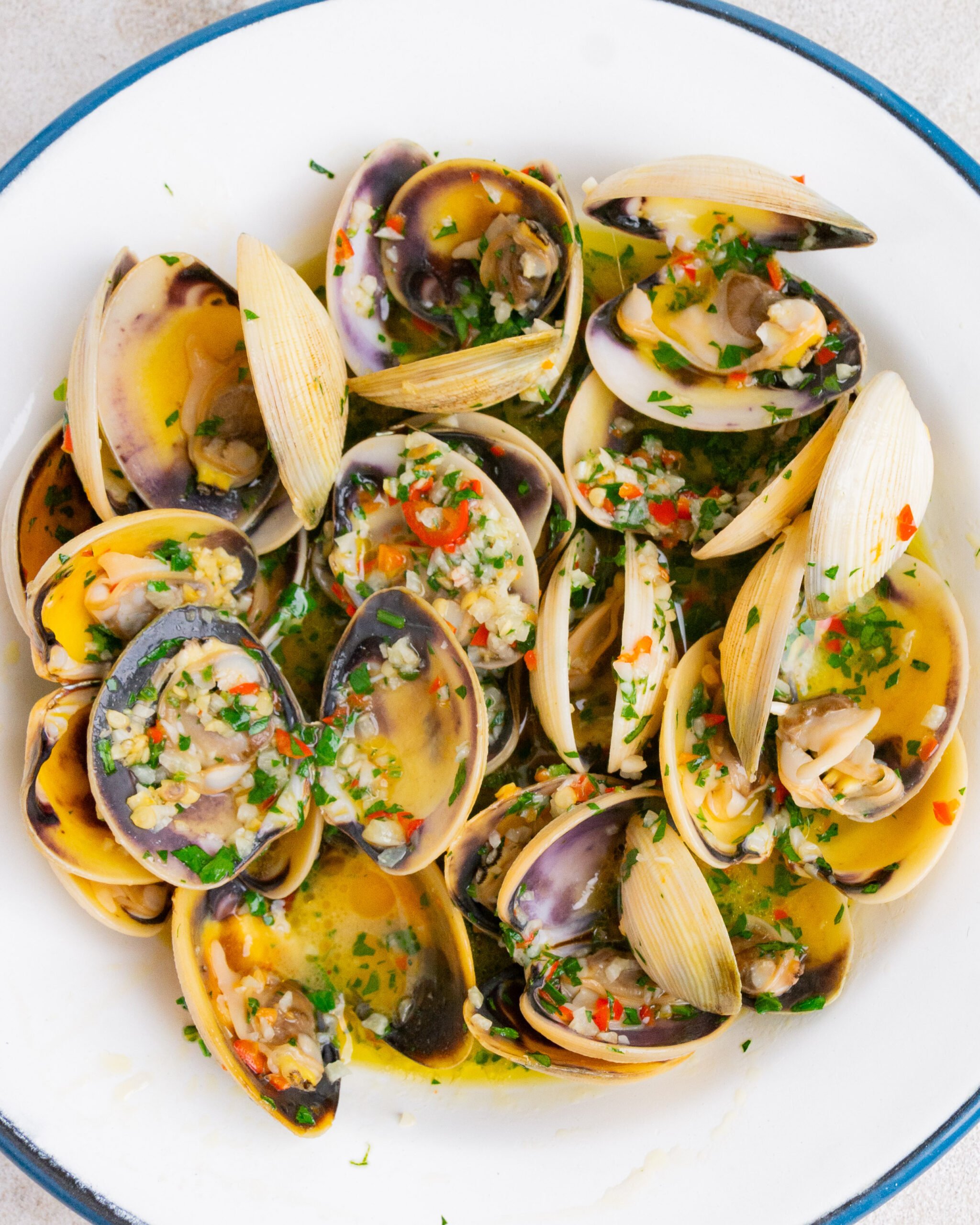 Garlic butter pipis / clams sit in large white bowl with. blue rim. They have parsley sprinkled over them and you can see lots of minced garlic and sliced chilli. 