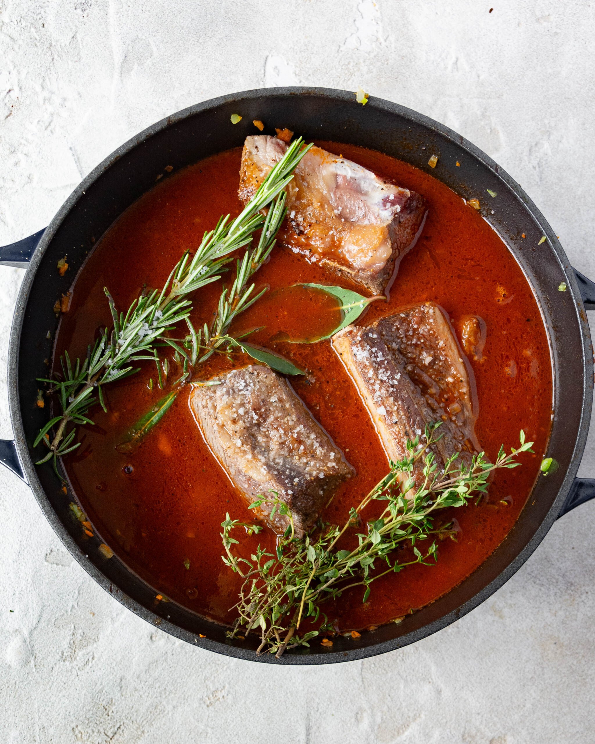 A big black pot is filled with a rich red sauce. You can see seared beef short ribs and fresh herbs sitting on top of the sauce. 