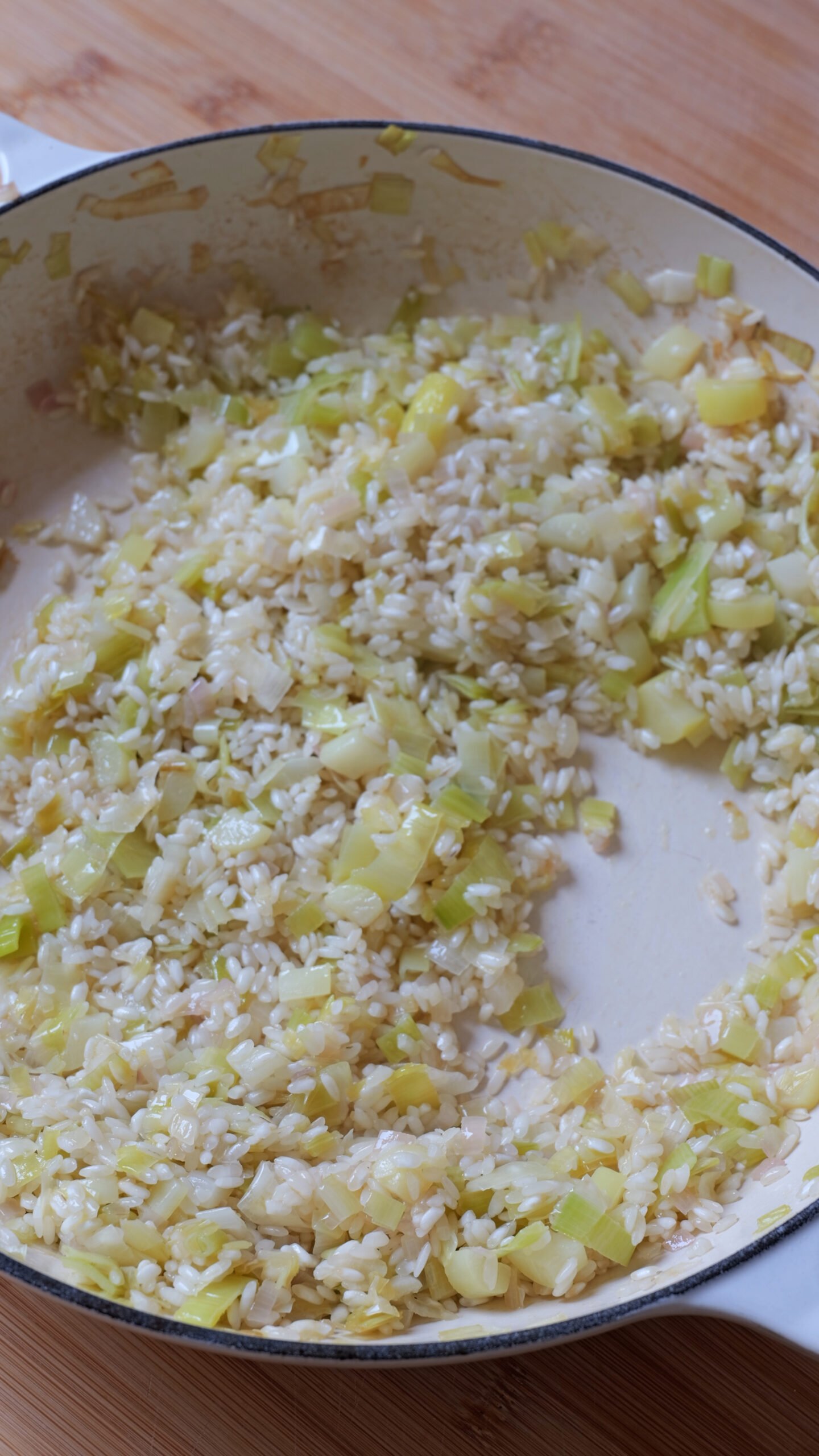 Leeks, shallot and garlic are frying in white pan with rice being toasted. 