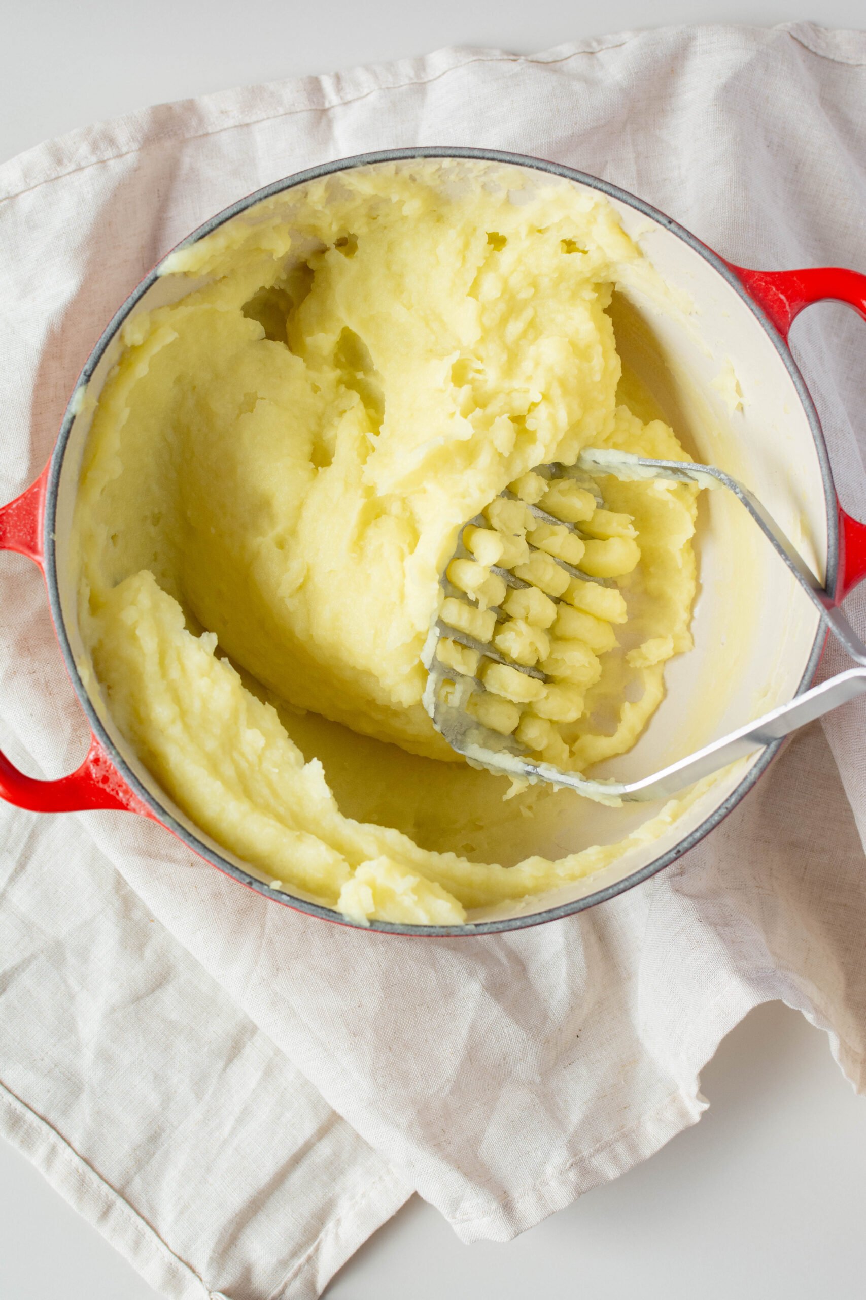 olive oil mashed potatoes in large red pot with potato masher sticking out of the side. 