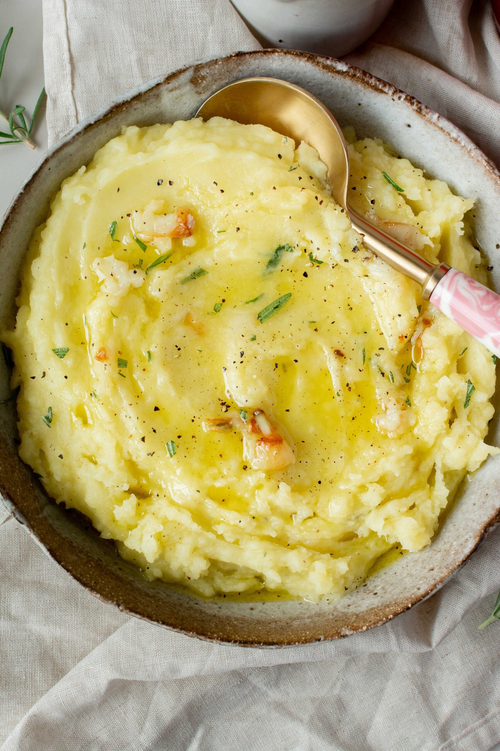 olive oil mashed potatoes in bowl. Seasoned with rosemary and black pepper. Gold spoon with pink handle is in the bowl. 