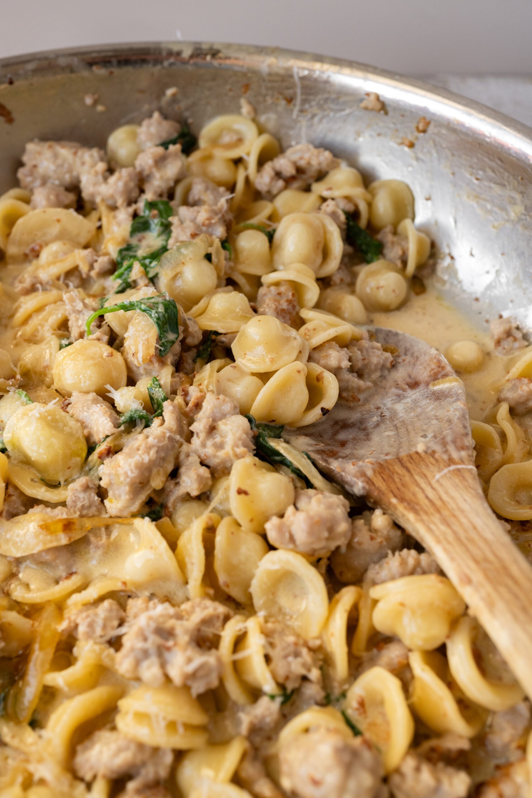 Caramelised Onion and Italian Sausage Orecchiette is shown in stainless steel pan with wooden spoon sticking out the side. 