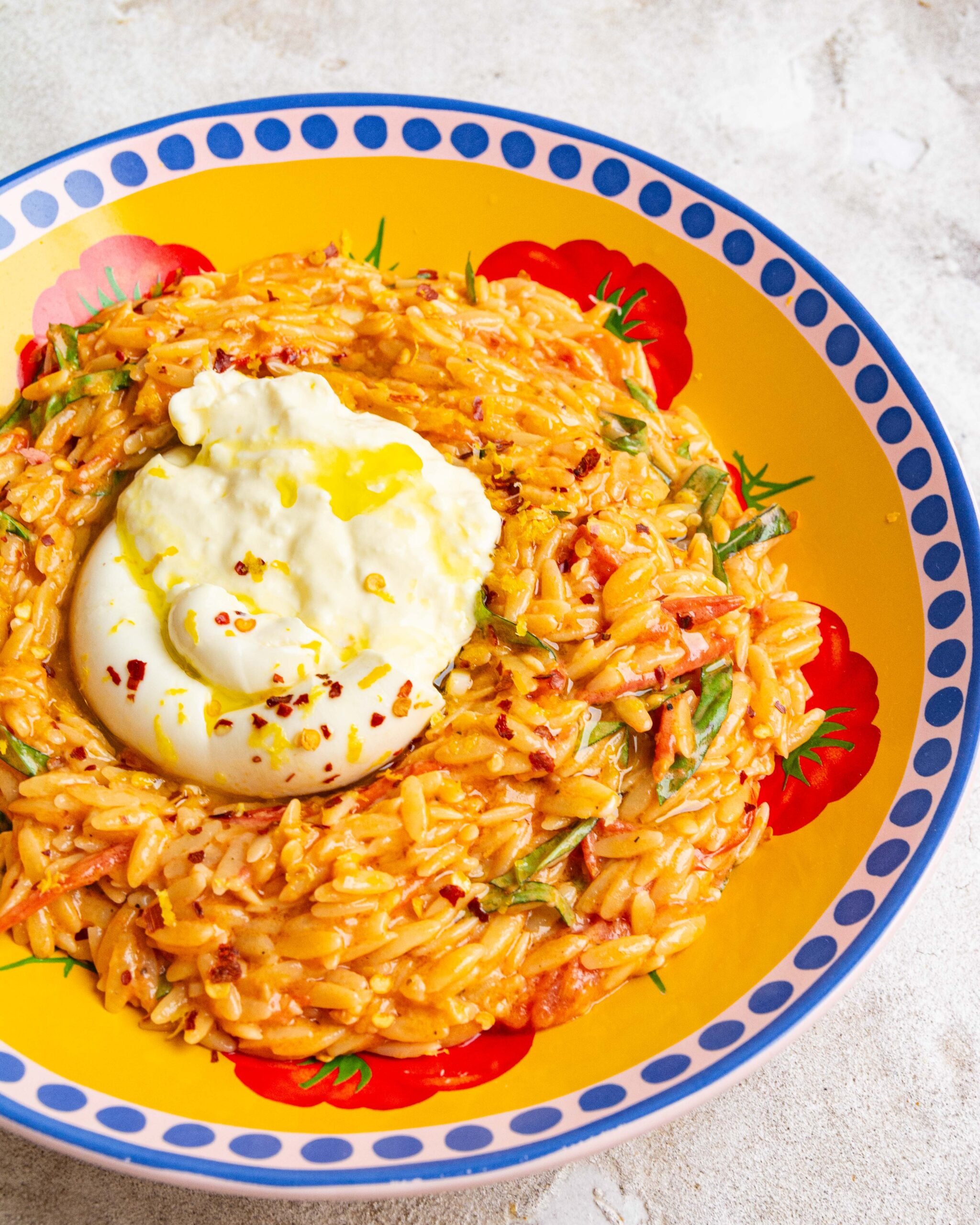 One pot creamy cherry tomato orzo sits in yellow bowl with blue dots around rim. Large burrata is on top and cracked open.