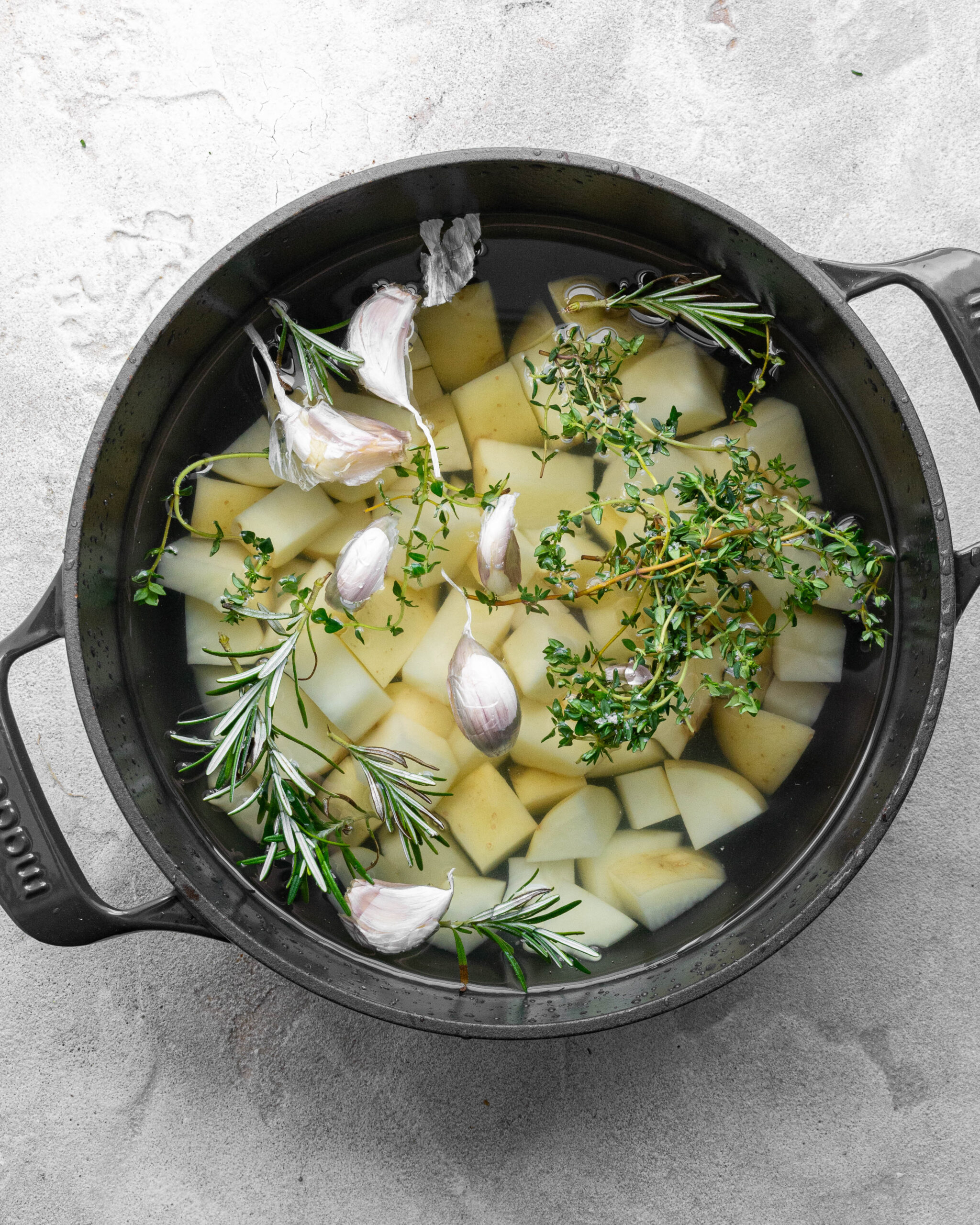 Sliced potatoes sit in large blue pot with garlic, sprigs of thyme and rosemary.
