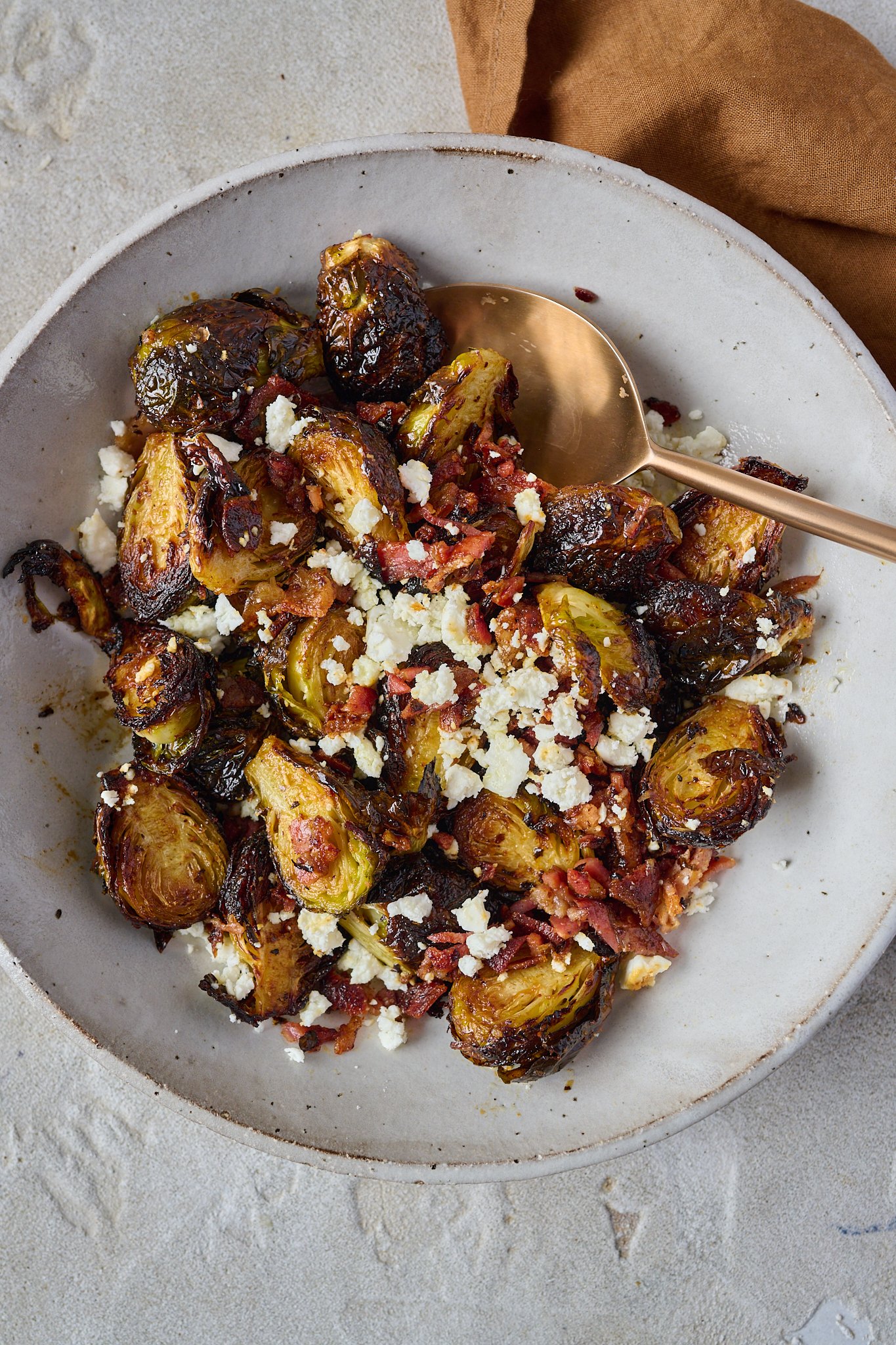 Crispy brussels sprouts sit in large white bowl with crispy bacon and feta crumbled on top. Gold spoon is sticking out of right side of bowl.