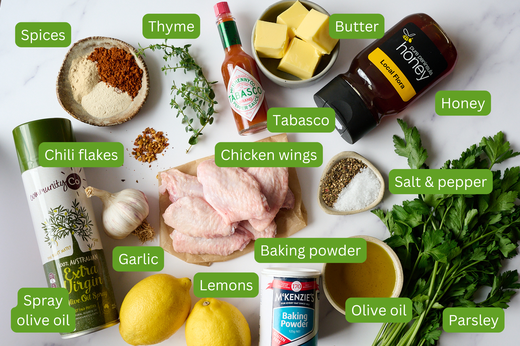 List of ingredients layed out on a board. Picture includes the items: spices, thyme, tabasco, butter, honey, chicken wings, chili flakes, salt and pepper, olive oil, parsley, baking powder, lemons and garlic. 