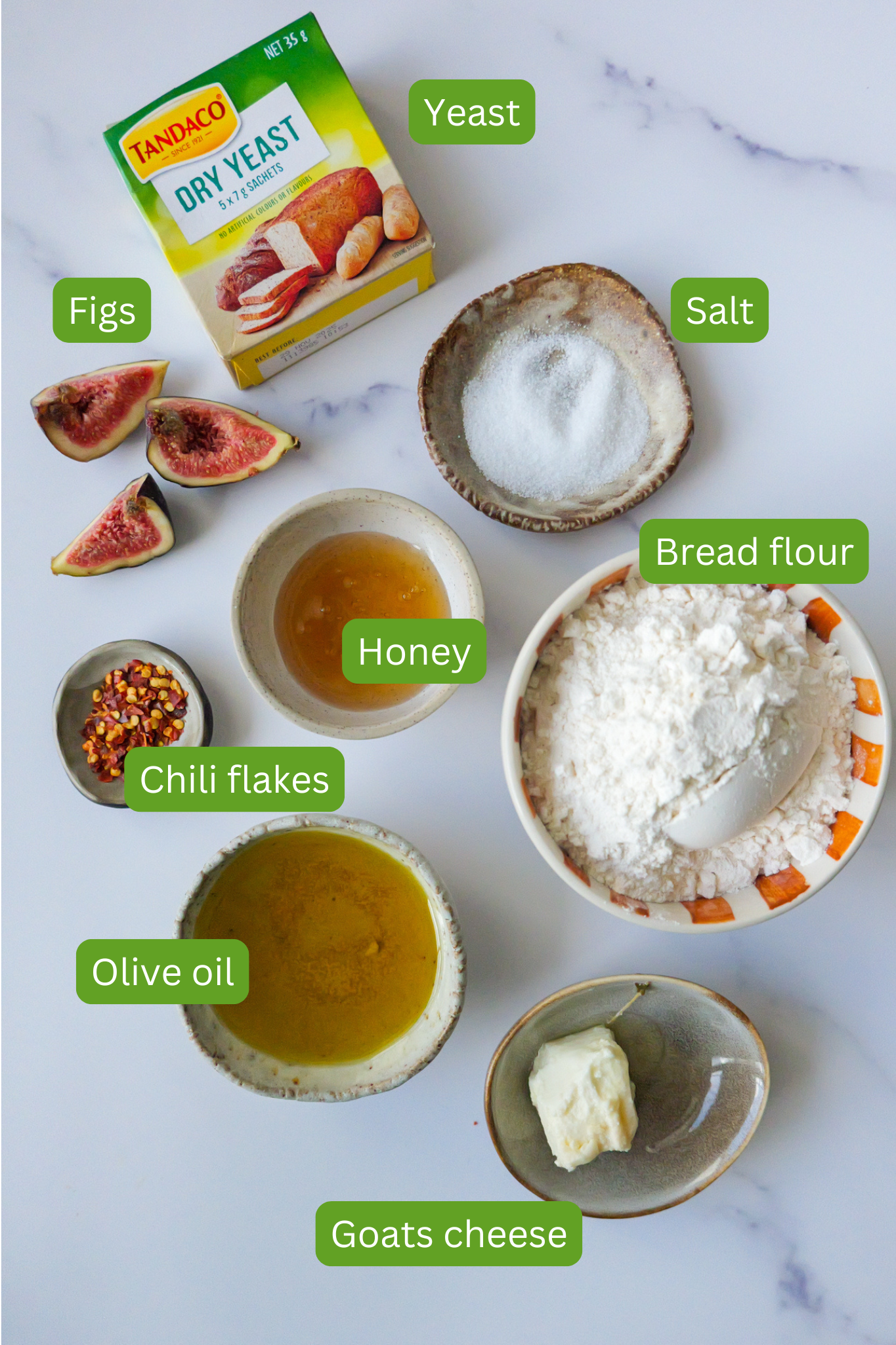 Ingredients for fig and goats cheese focaccia are laid out on marble countertop. Ingredients include olive oil, goats cheese, chilli flakes, flour, honey, figs, salt and yeast
