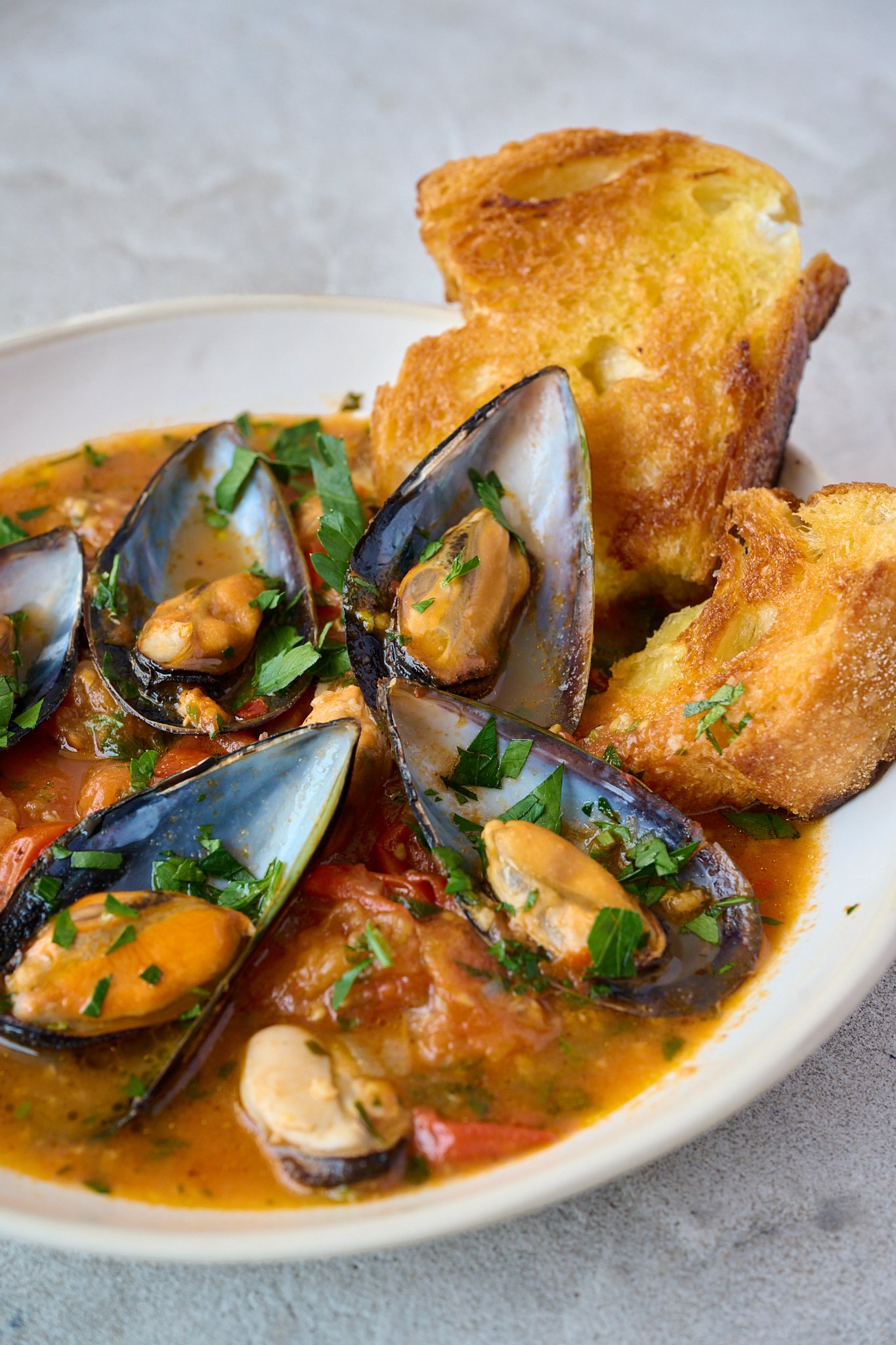 Mussels and tomato sauce served in a white bowl with fresh parsley sprinkled over the top and white sourdough on side of bowl. 
