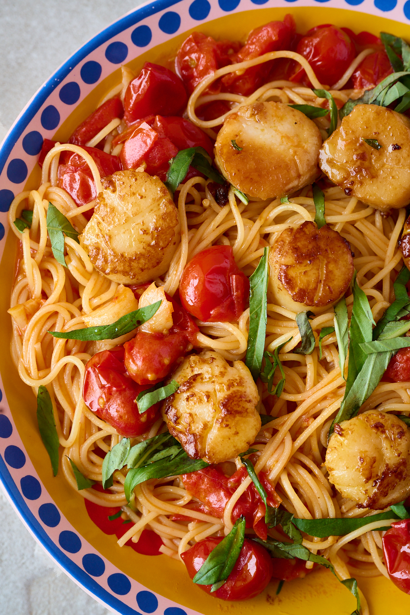 perfectly seared scallops resting on a bed of cherry tomato pasta in a bowl with blue dots around the rim. A sprinkling of fresh basil over the top of the pasta. 