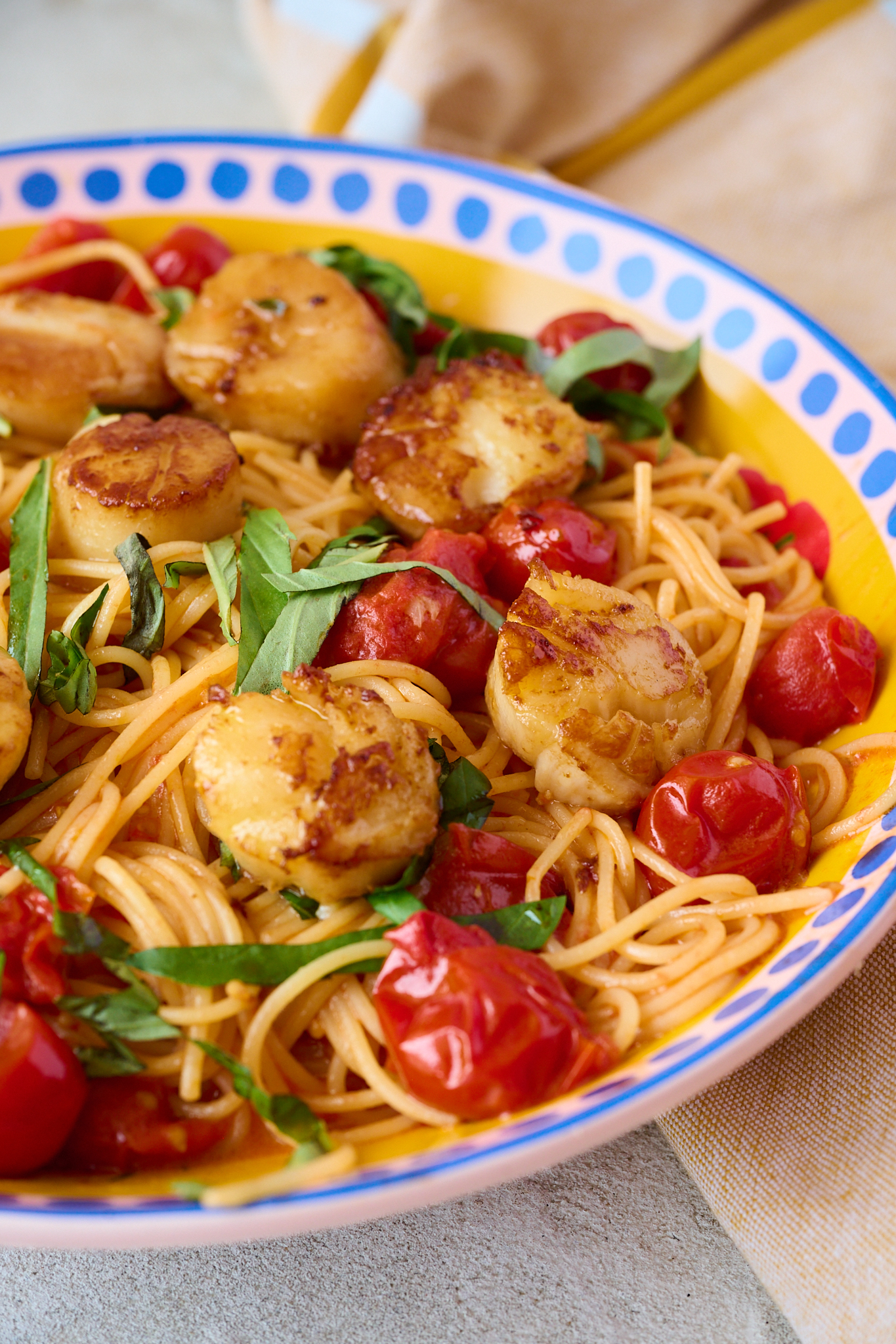 perfectly seared scallops resting on a bed of cherry tomato pasta in a bowl with blue dots around the rim. A sprinkling of fresh basil over the top of the pasta. 