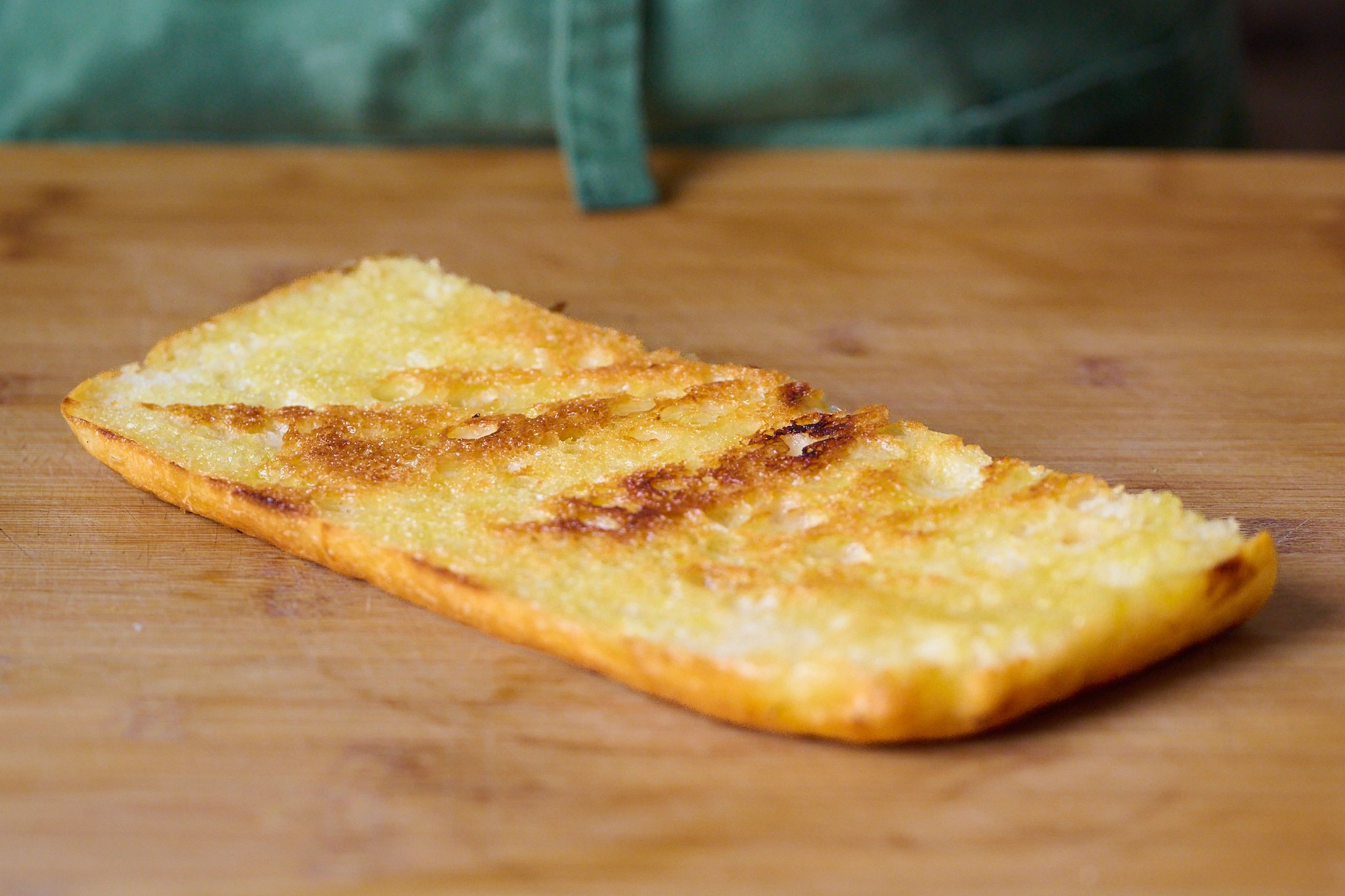 Piece of focaccia that has been slightly toasted. Sits on wooden chopping board. 