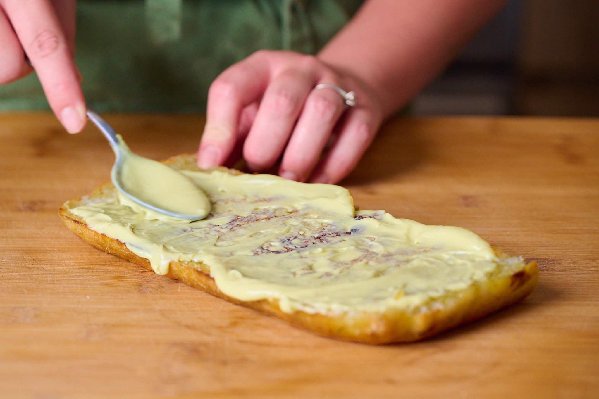 Piece of focaccia that has been slightly toasted and now has a layer of garlic confit aioli. Sits on wooden chopping board. 
