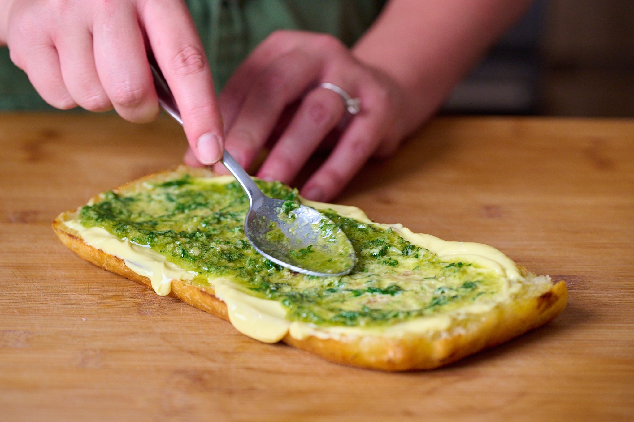 Piece of focaccia that has been slightly toasted and now has a layer of garlic confit aioli and pesto. Sits on wooden chopping board. 