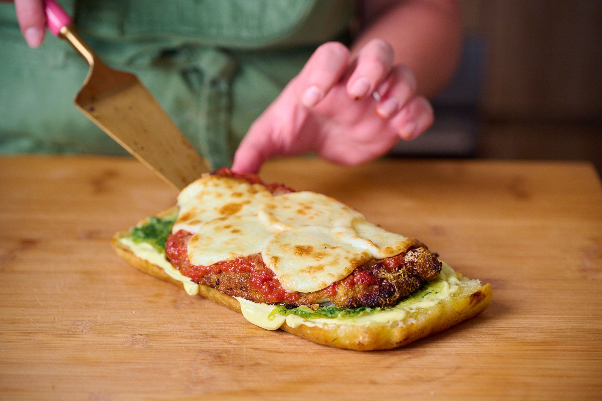Piece of focaccia that has been slightly toasted and now has a layer of garlic confit aioli, pesto and the chicken parm being put on top. Sits on wooden chopping board. 