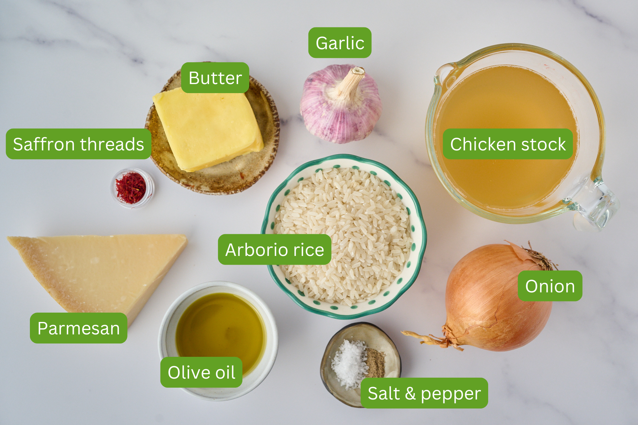 List of ingredients laid out on white background for risotto alla milanese. Ingredients include parmesan, olive oil, salt and pepper, onion, arborio rice, butter, saffron threads, garlic and chicken stock. 