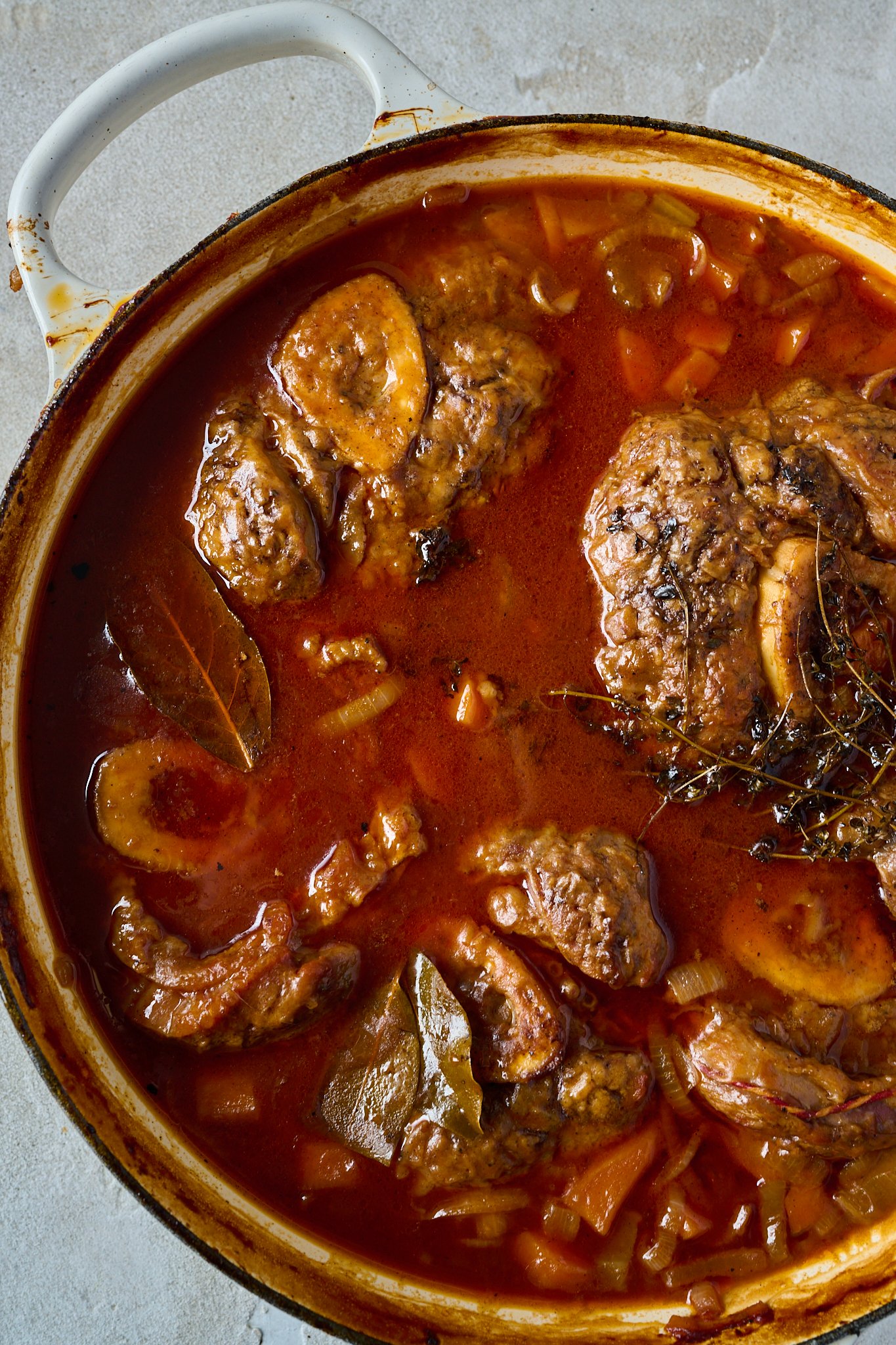 Osso buco has been made is in large white pan with a blue rim. The sauce is deep red and you can see the veal shanks and herbs. 