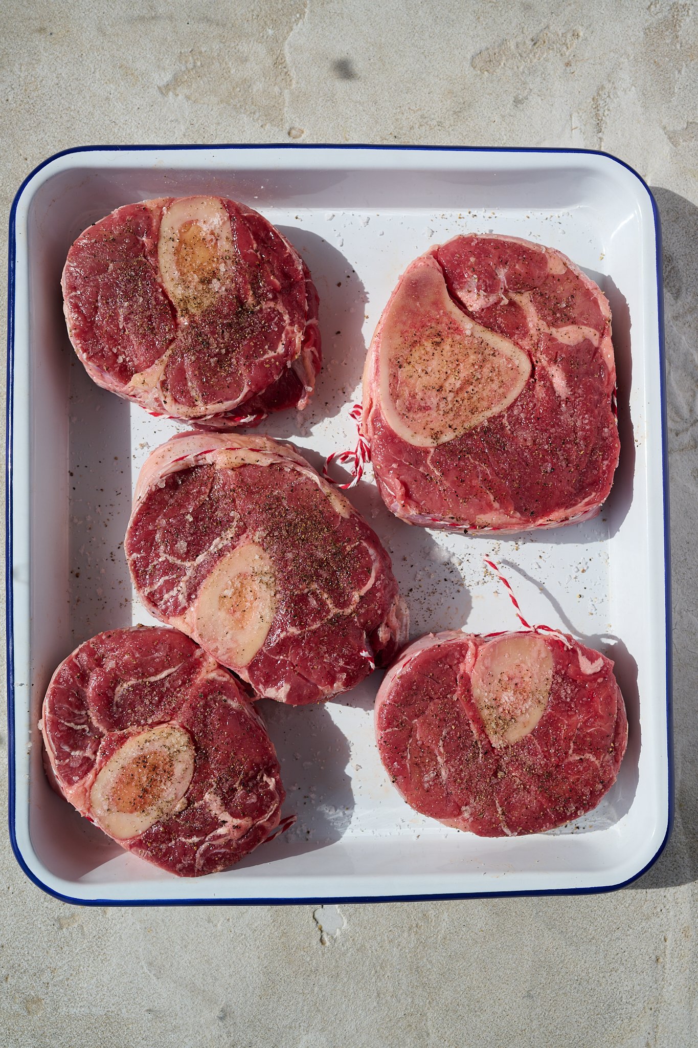 Veal shanks sit in white baking tray. They are seasoned with salt and pepper and have red and white butchers string around them. 