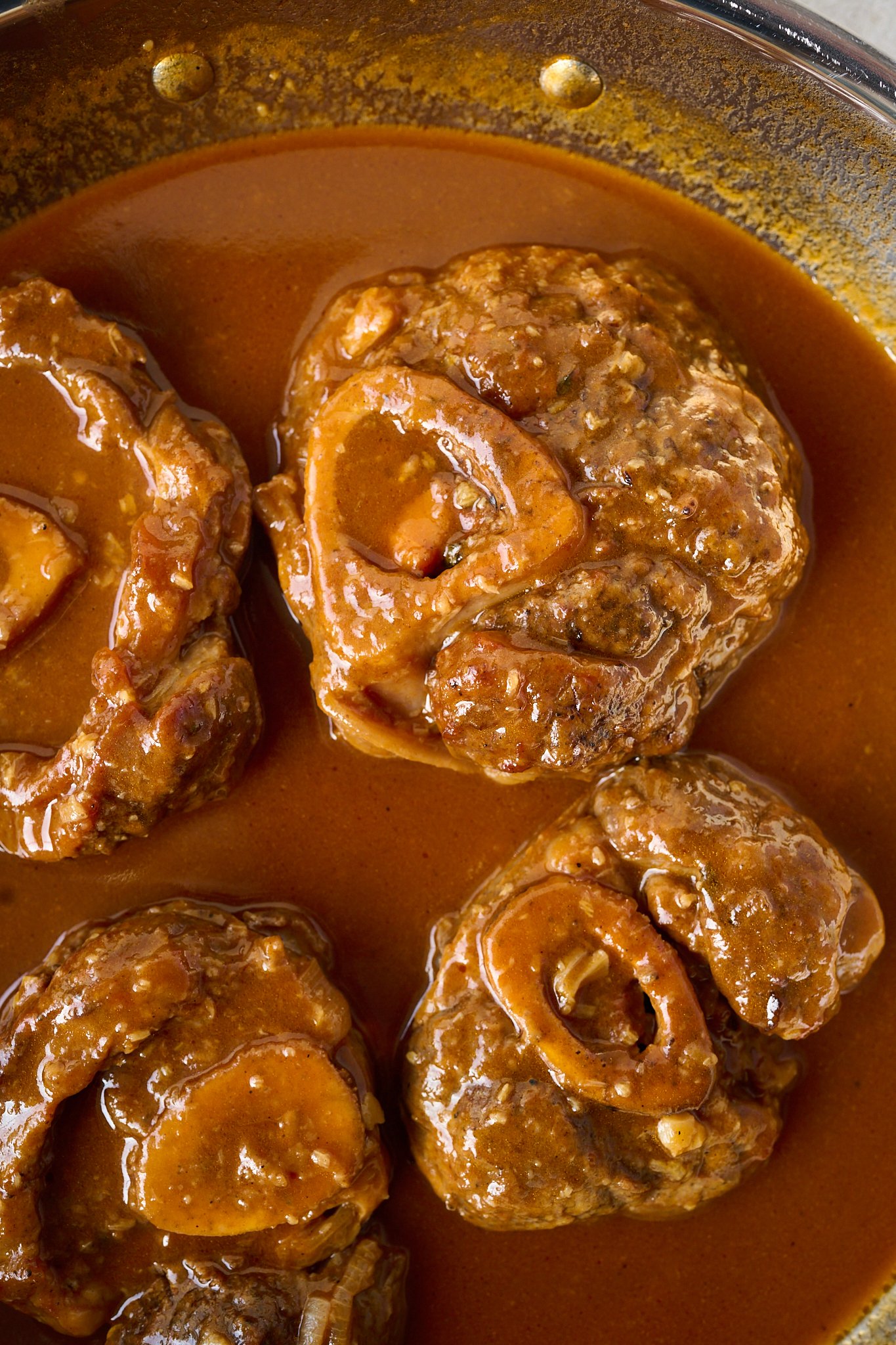 veal shanks are basted in strained red sauce and sit in a metal pan. 