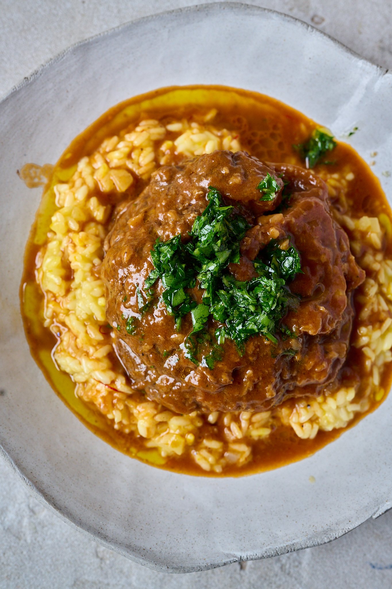 Osso buco sits on a bed of saffron risotto in a large white bowl with gremolata spooned on top. lots of sauce has pooled at the bottom.